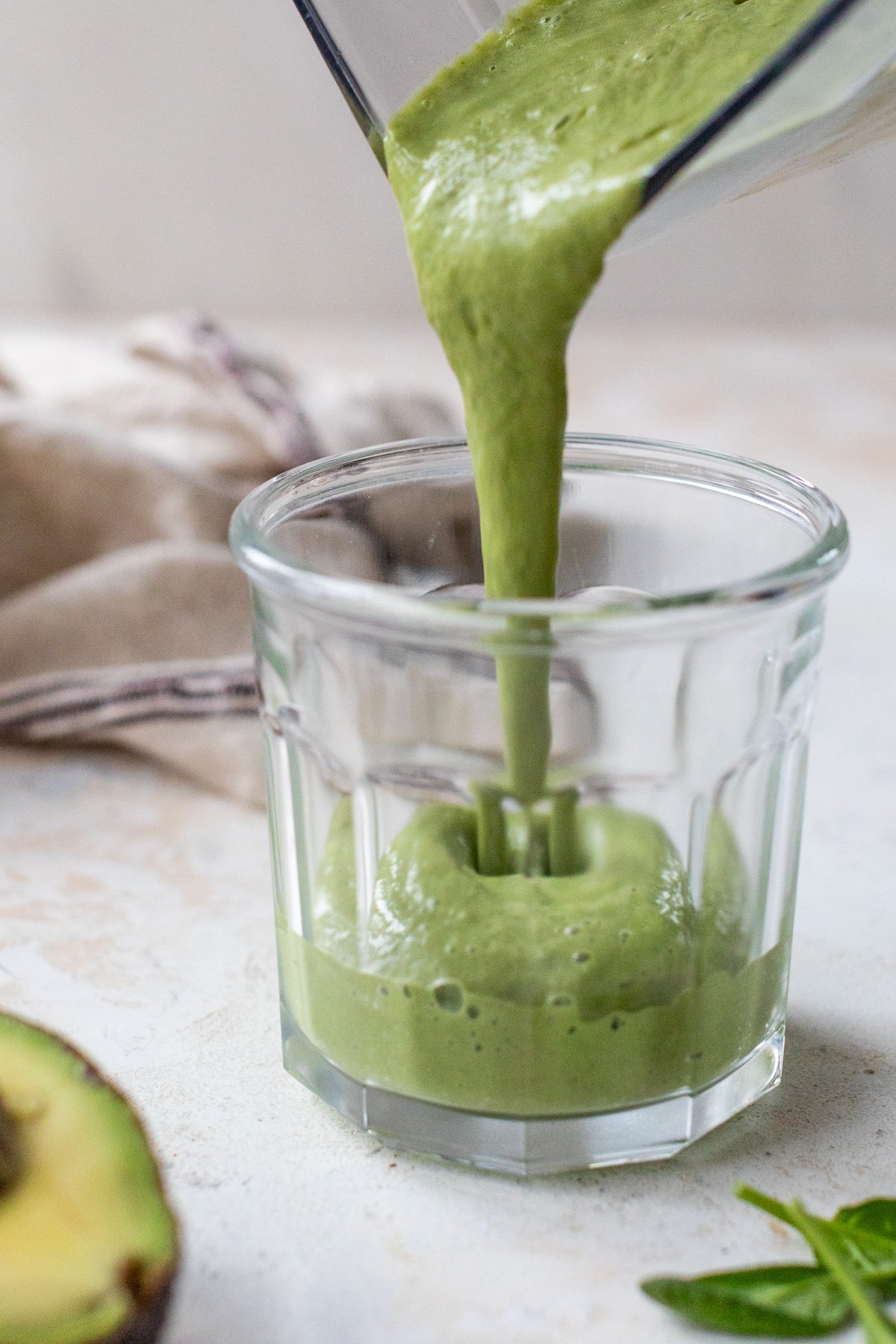 A thick green avocado smoothie is being poured from a blender into a clear glass. An avocado half and some spinach leaves lie nearby on a light surface. A tan cloth with stripes is in the background.
