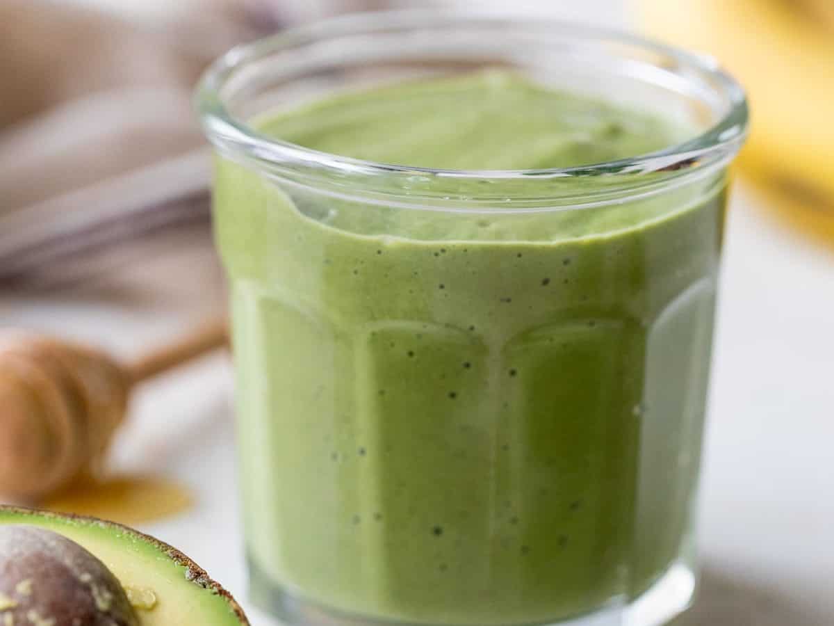 A glass filled with a thick avocado smoothie sits on a light surface, surrounded by a halved avocado, fresh spinach leaves, a honey dipper, and a banana in the background.