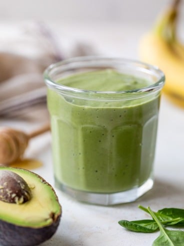 A glass filled with a thick avocado smoothie sits on a light surface, surrounded by a halved avocado, fresh spinach leaves, a honey dipper, and a banana in the background.