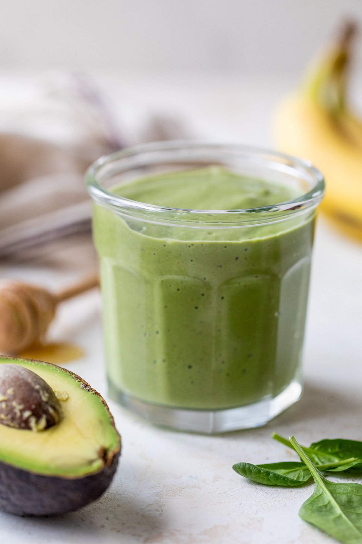 A glass filled with a thick avocado smoothie sits on a light surface, surrounded by a halved avocado, fresh spinach leaves, a honey dipper, and a banana in the background.