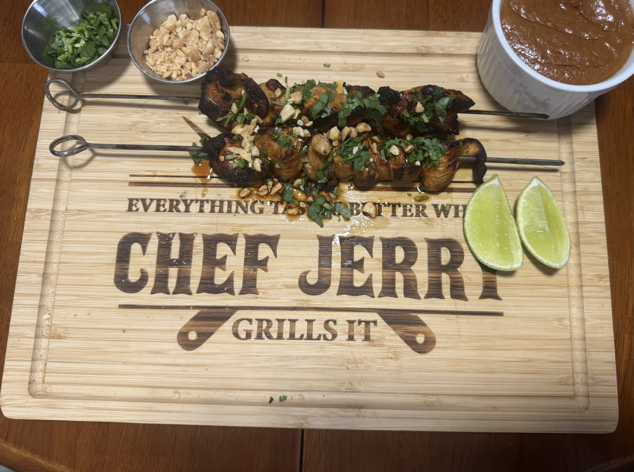 Two chicken skewers with chopped peanuts and herbs on a cutting board, with lime wedges, sauce, and toppings on the side.