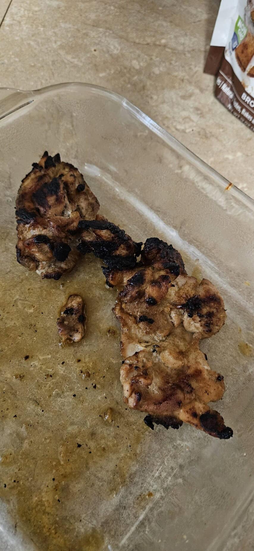 Grilled pieces of chicken rest on a glass baking dish with some oil and grill marks visible.