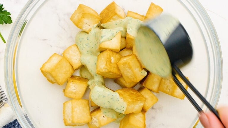 tossing roasted crispy tofu with tahini dressing in a mixing bowl for buddha bowls