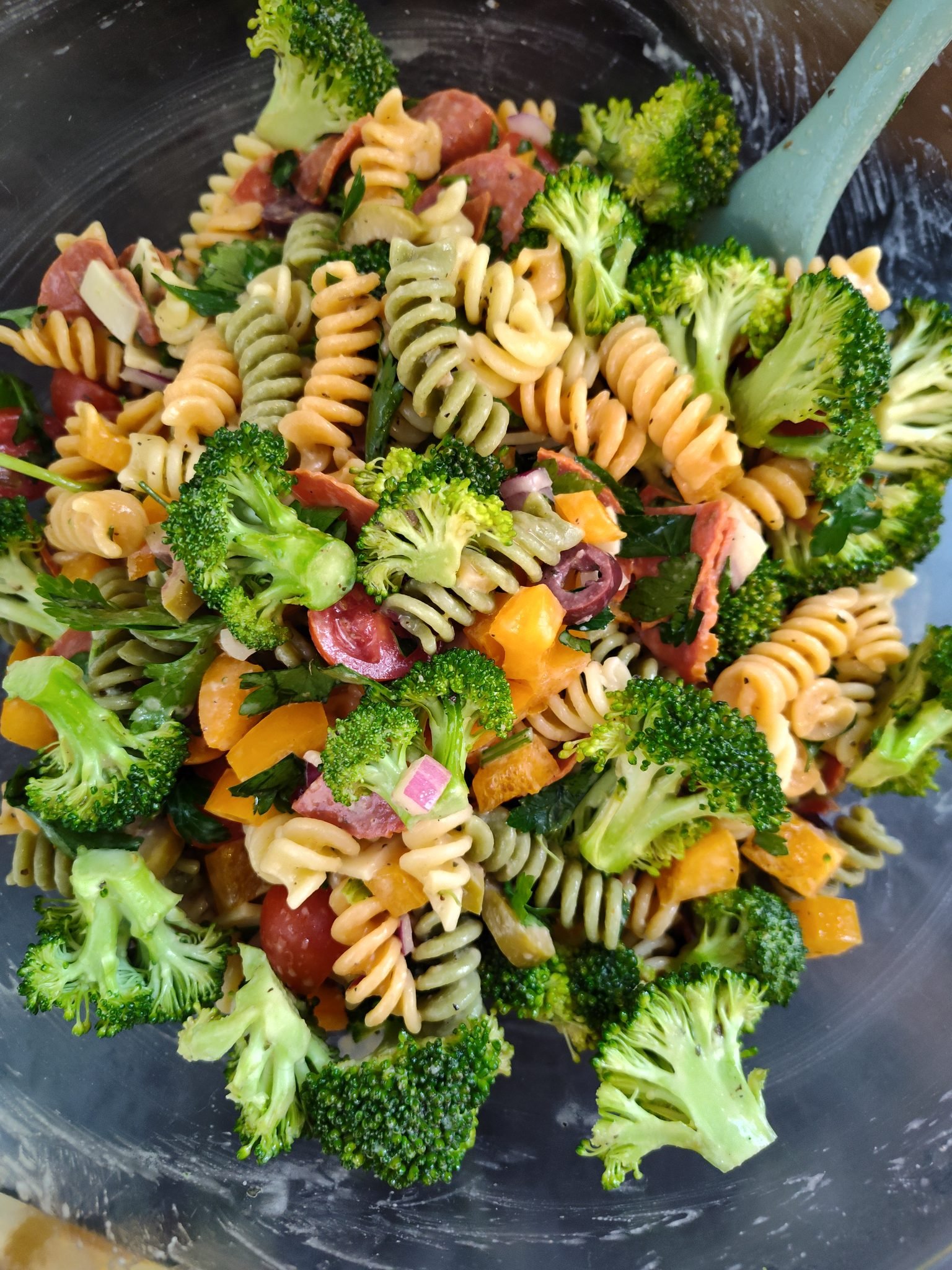 A colorful pasta salad with rotini noodles, fresh broccoli florets, chopped yellow bell peppers, tomatoes, red onions, and olives, mixed together in a bowl.