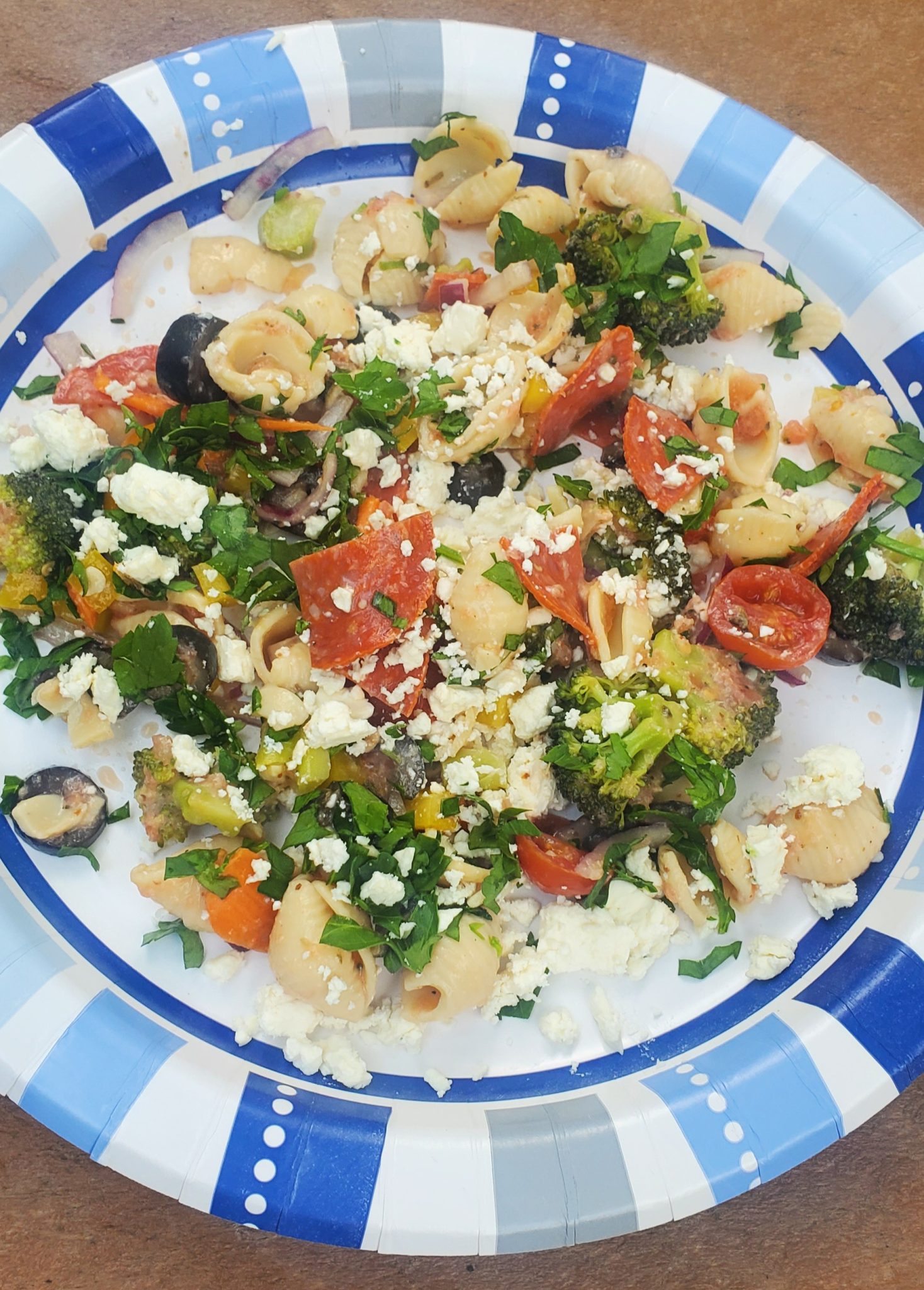 Pasta salad with vegetables, pepperoni, olives, and crumbled cheese on a blue and white paper plate.