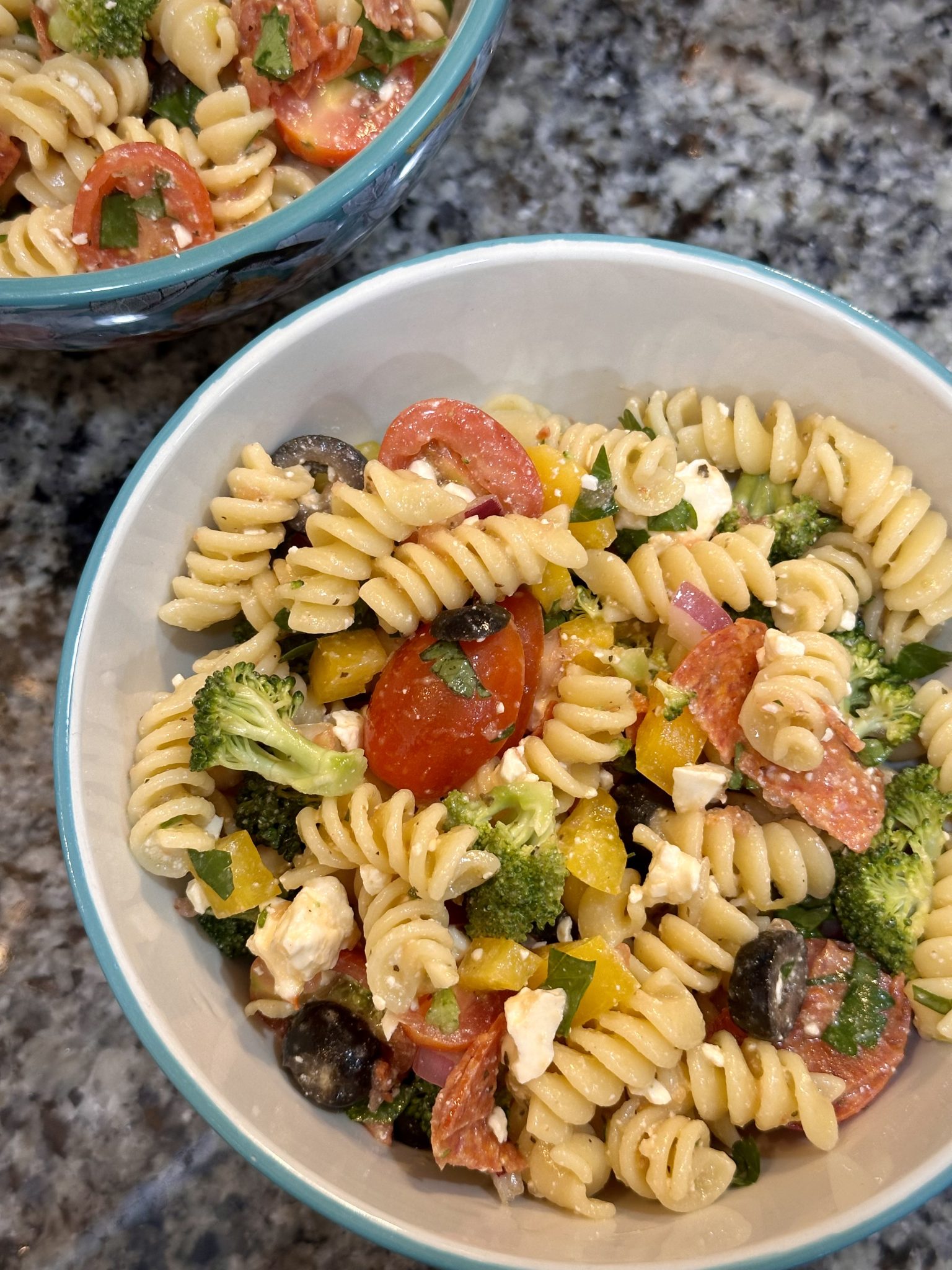 A bowl of pasta salad with rotini noodles, cherry tomatoes, broccoli, yellow bell peppers, black olives, red onion, feta cheese, pepperoni slices, and chopped herbs on a granite countertop.