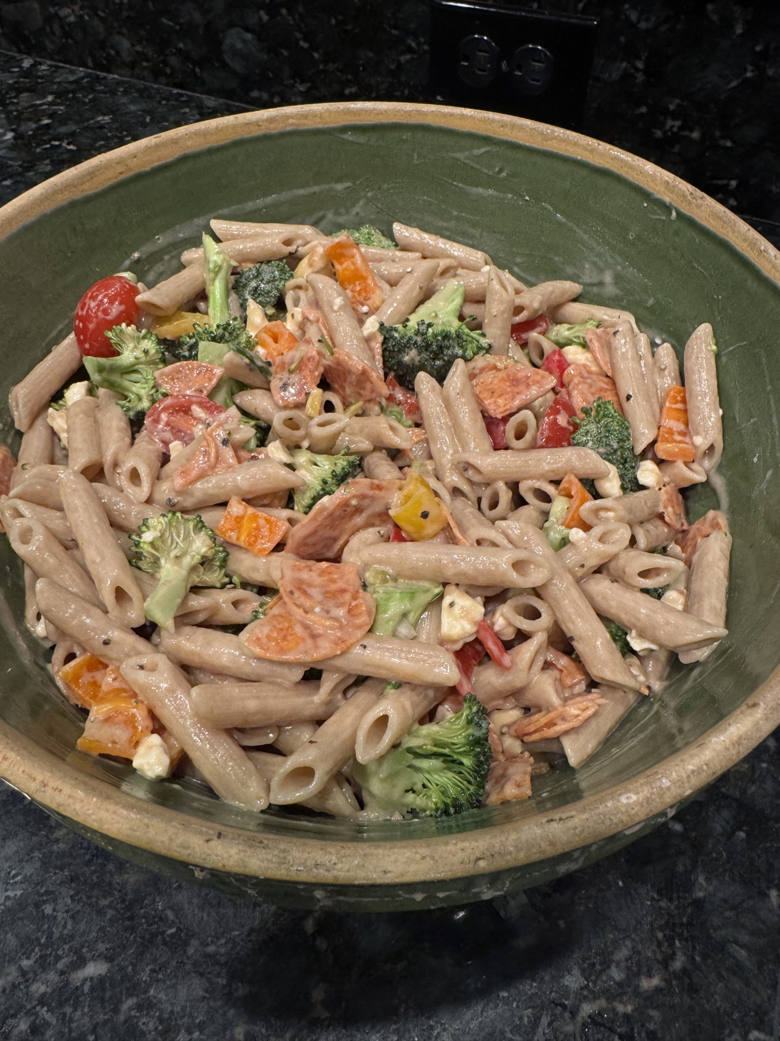 A green bowl filled with whole wheat penne pasta mixed with broccoli, cherry tomatoes, orange bell peppers, and other assorted vegetables in a creamy sauce on a dark countertop.