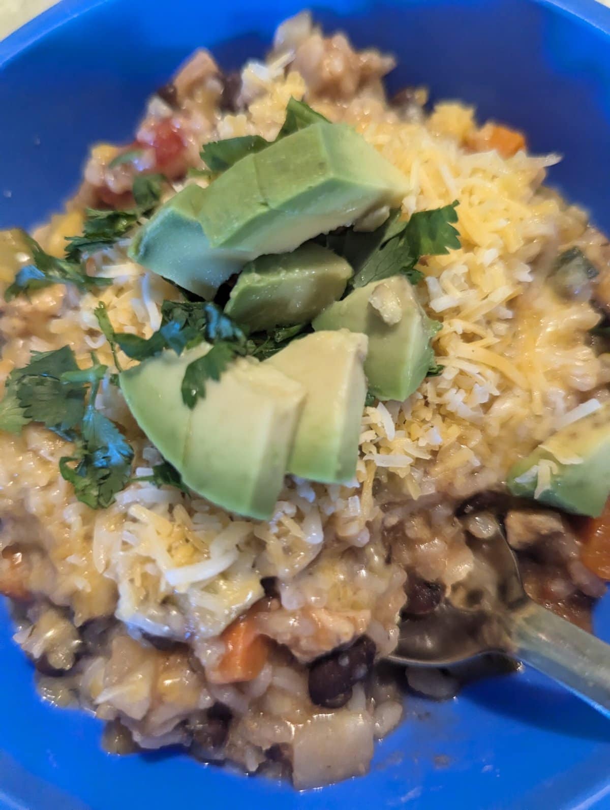 A bowl of rice and vegetable stew topped with shredded cheese, cilantro, and avocado chunks, with a spoon inside.