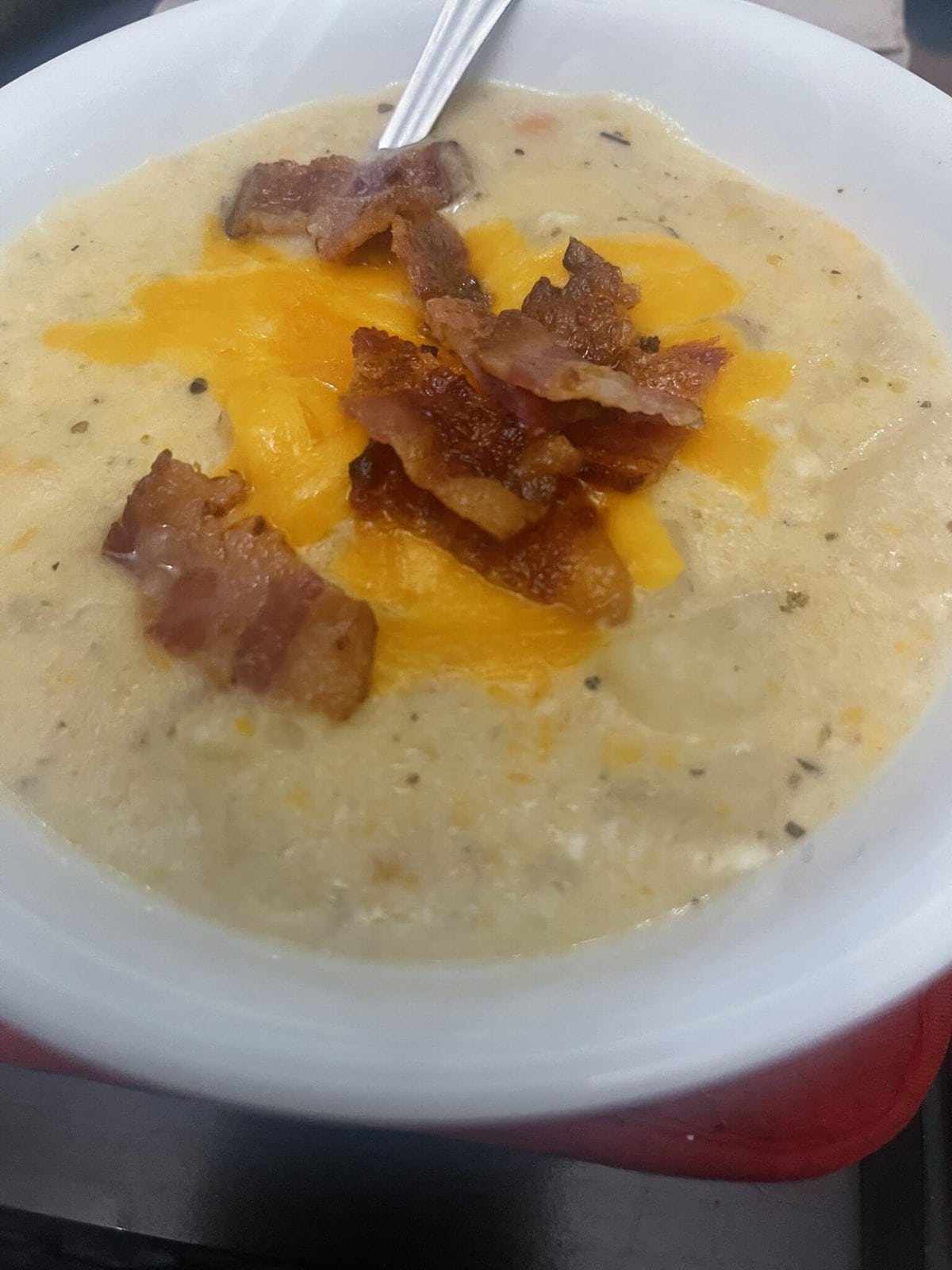 A bowl of creamy soup topped with shredded cheddar cheese and crispy bacon strips, with a spoon inside.