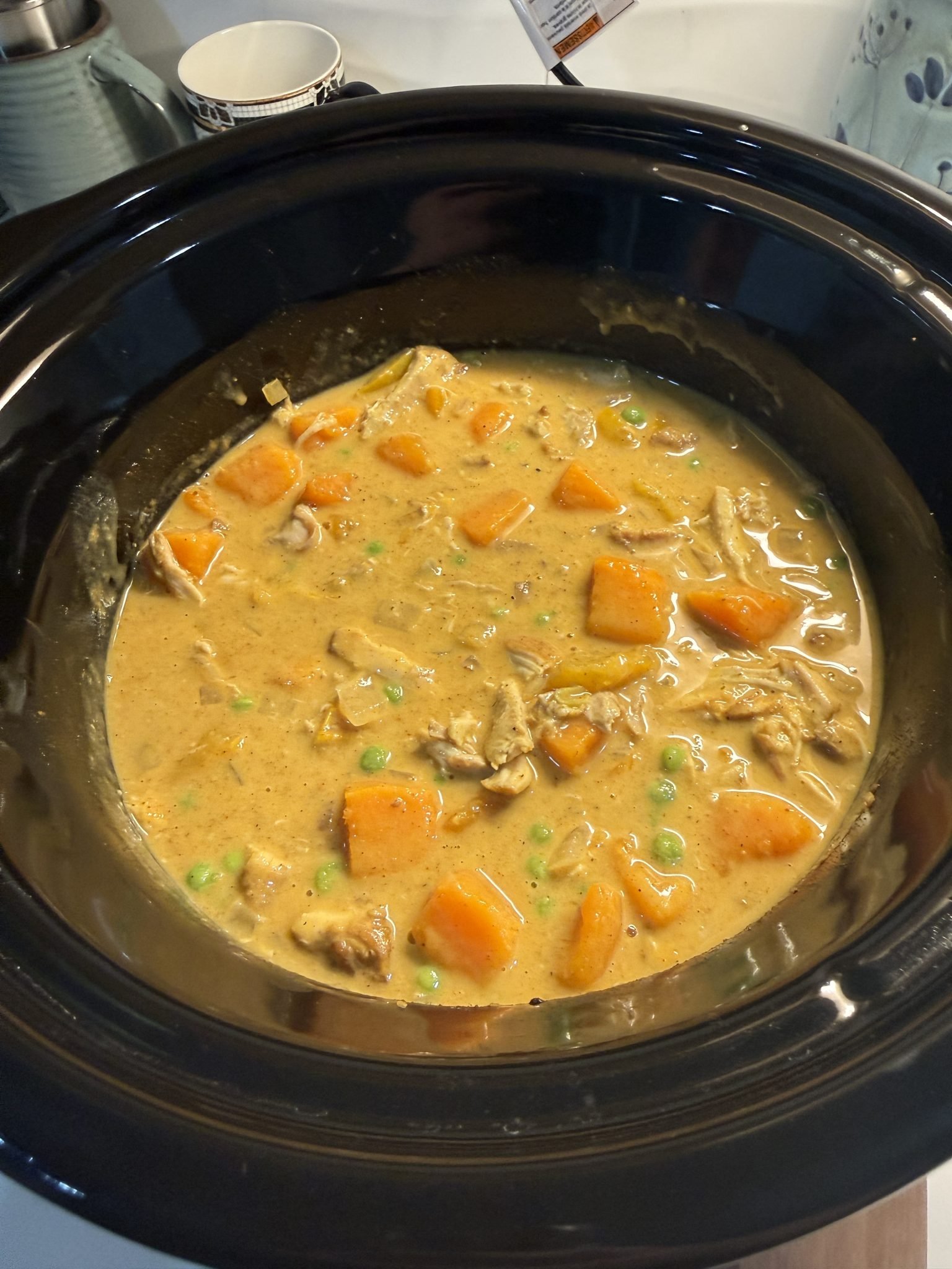 Chicken stew with carrots and peas simmering in a creamy sauce inside a black slow cooker.