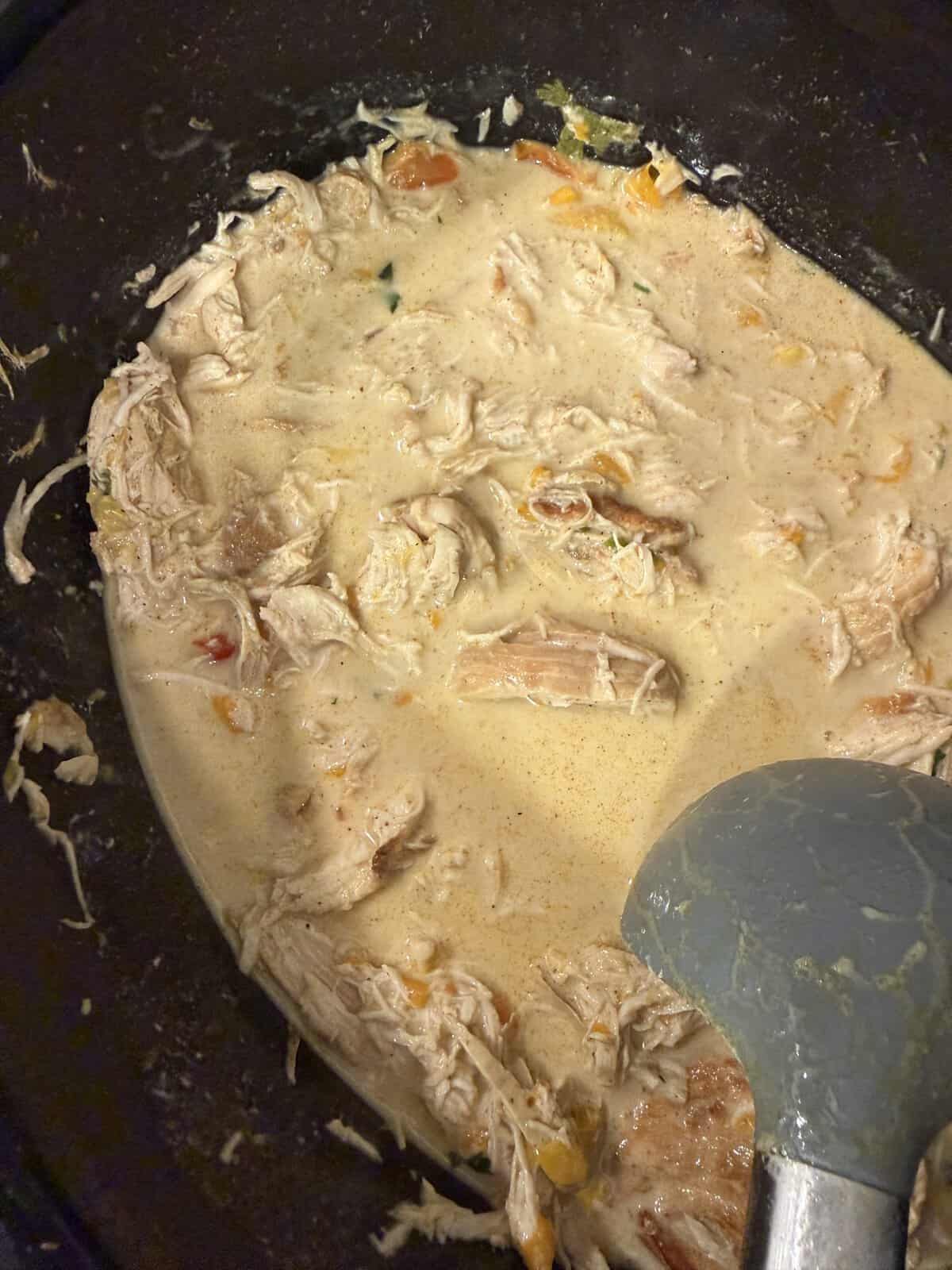 Creamy shredded chicken with vegetables in a slow cooker, stirred with a gray ladle.