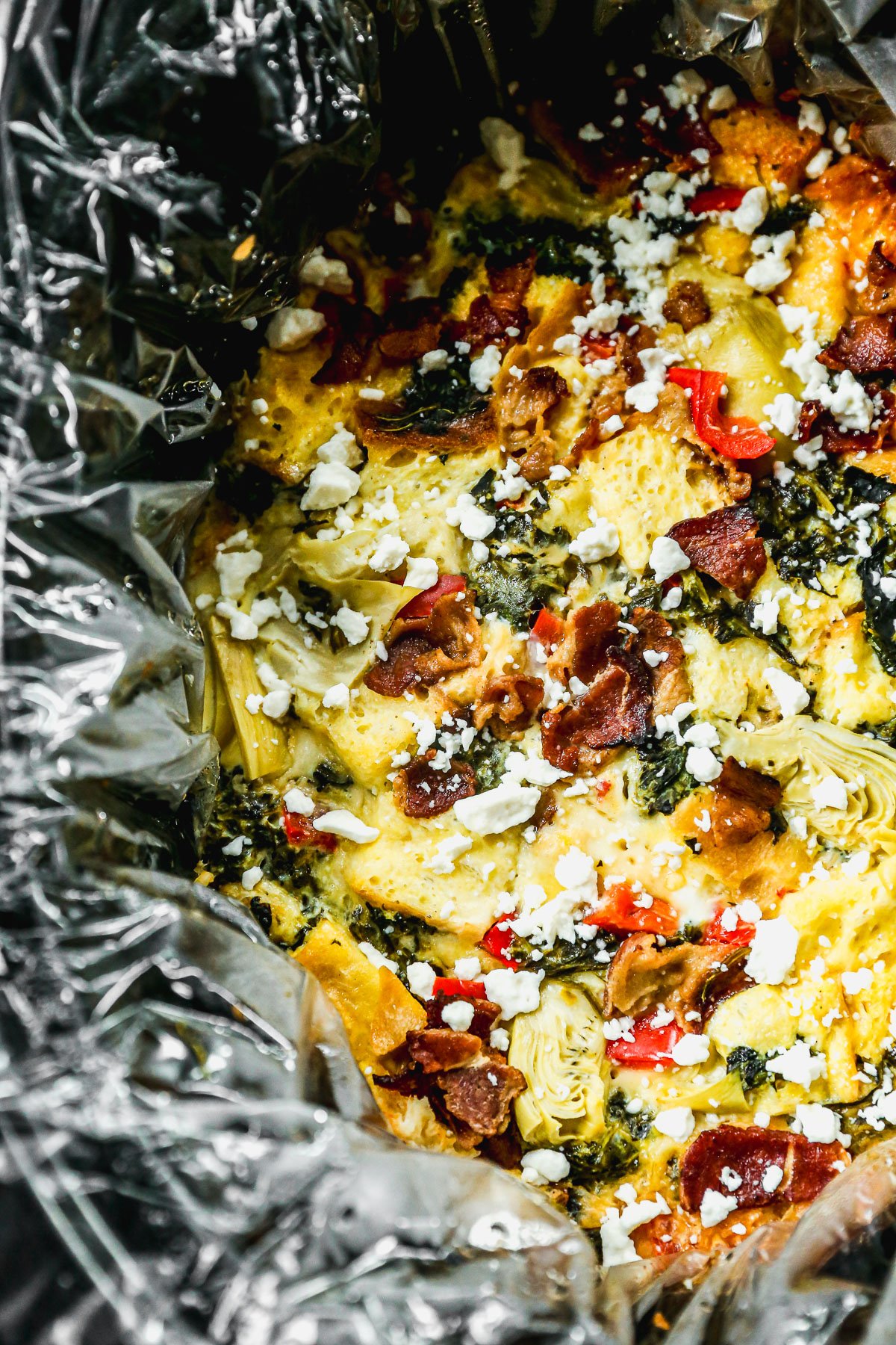 Breakfast casserole with bacon, artichokes, spinach, and feta cheese in a foil-lined dish.