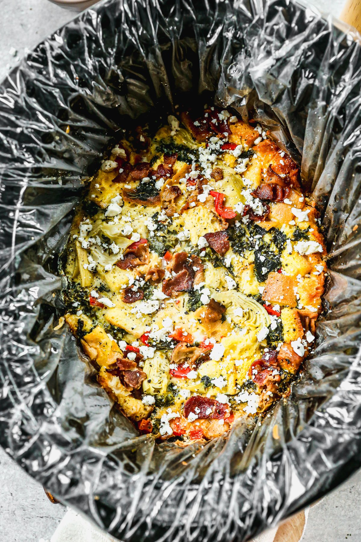 Slow cooker lined with a plastic liner filled with breakfast casserole topped with cheese, vegetables, and herbs.