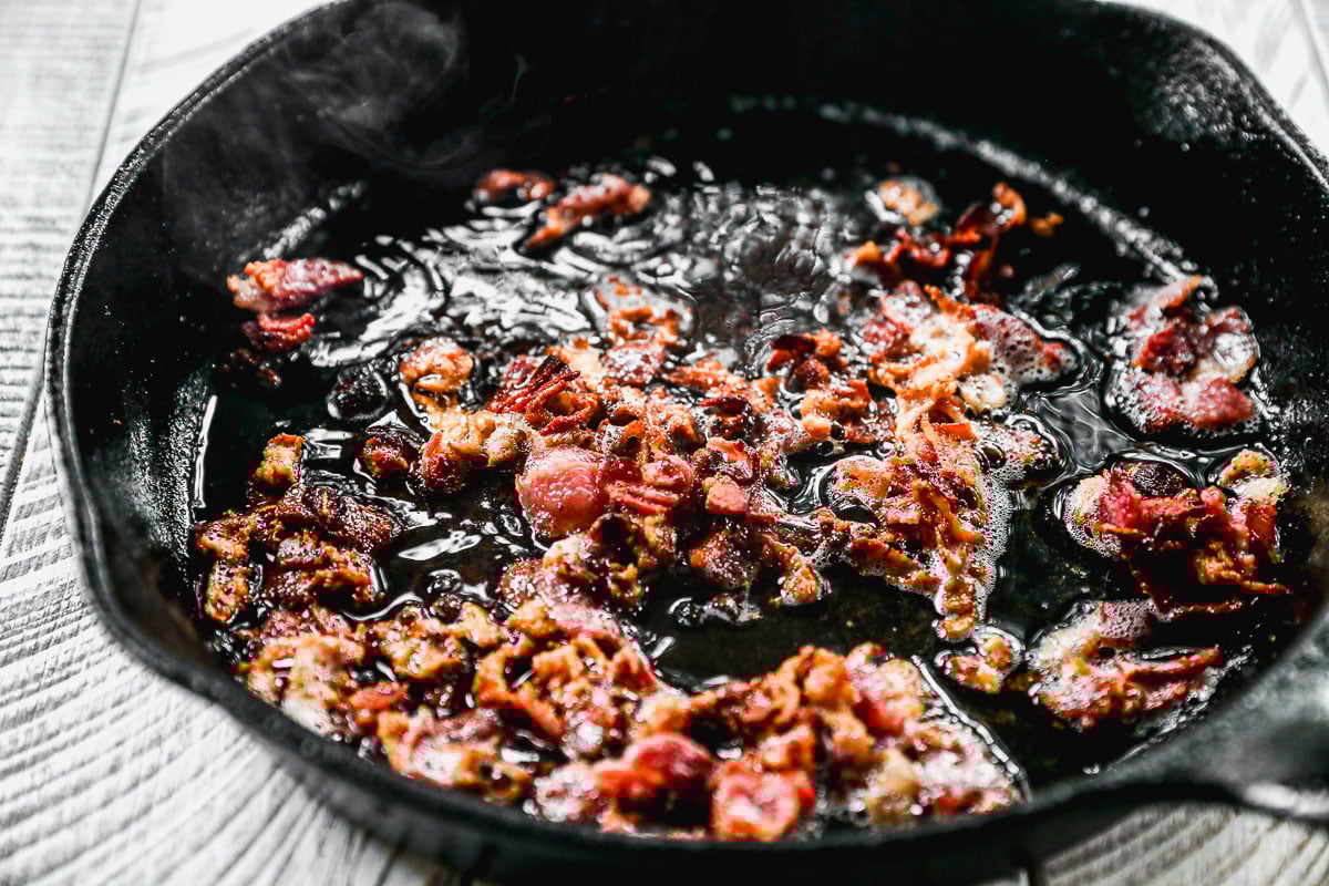 Crispy bacon pieces sizzling in hot grease inside a black cast iron skillet on a light wooden surface.
