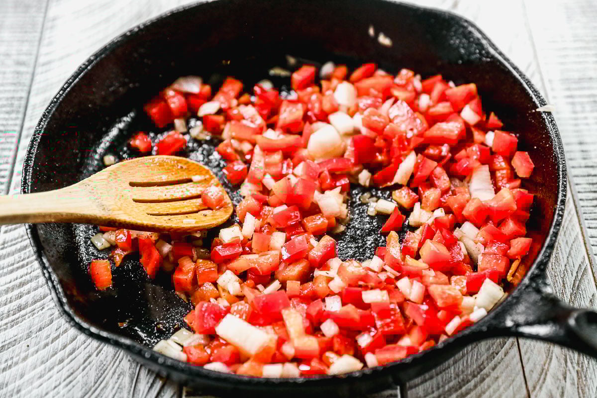 Chopped red bell peppers and onions cooking in a black skillet with a wooden spoon on a wooden surface.