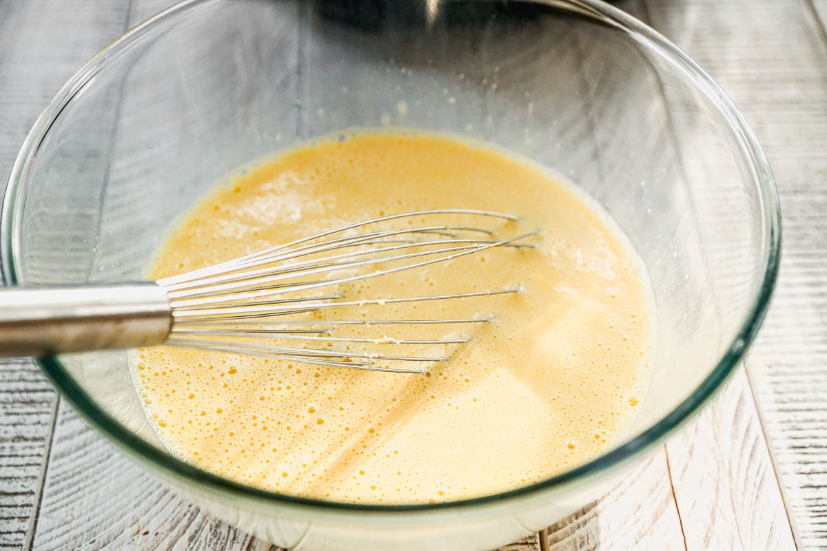 A metal whisk mixing beaten eggs in a clear glass bowl on a light wooden surface.