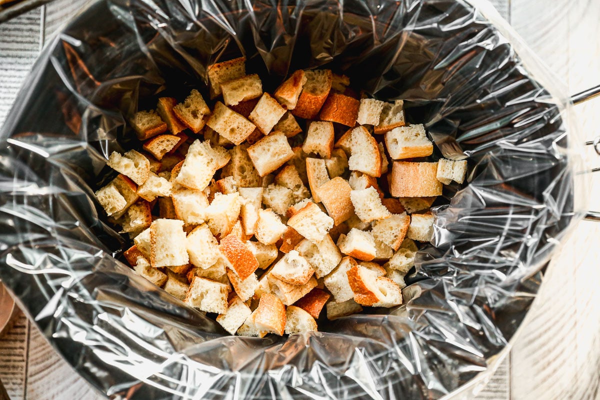 Cubed bread pieces in a slow cooker lined with a clear plastic liner, ready for cooking.