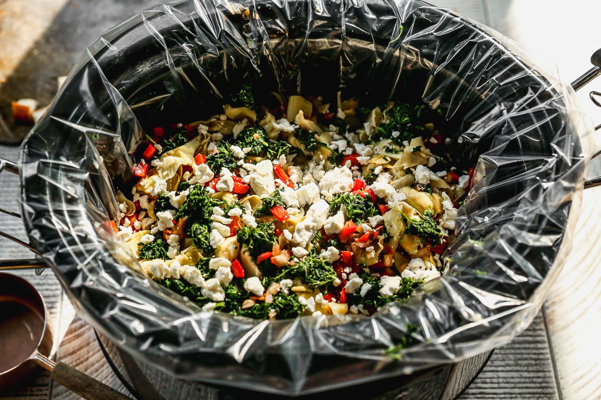 A slow cooker lined with plastic, filled with colorful chopped vegetables, greens, and crumbled cheese.
