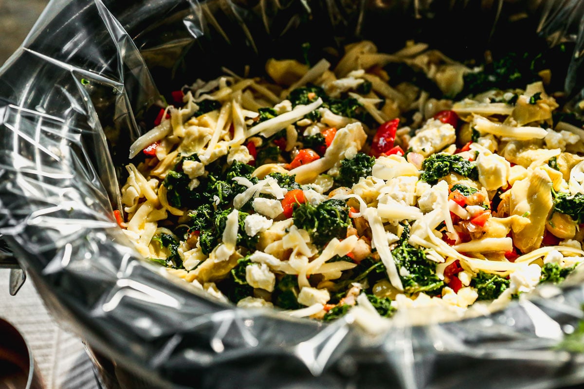 A slow cooker filled with a mixture of chopped vegetables, cheese, and greens lined with a plastic liner.