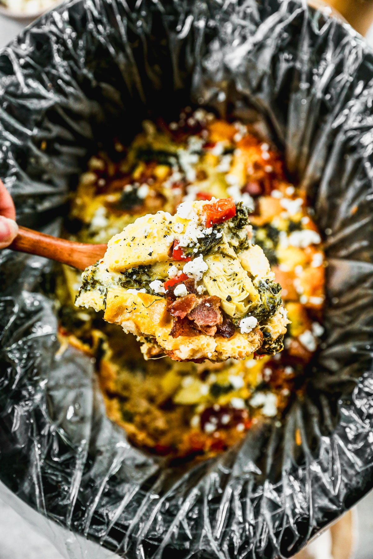 A spoon scoops a cheesy crockpot breakfast casserole with vegetables and sauce from a slow cooker lined with a plastic liner.