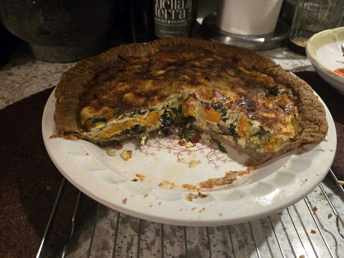 A baked quiche with a slice missing, showing a filling of vegetables on a decorative plate on a kitchen counter.