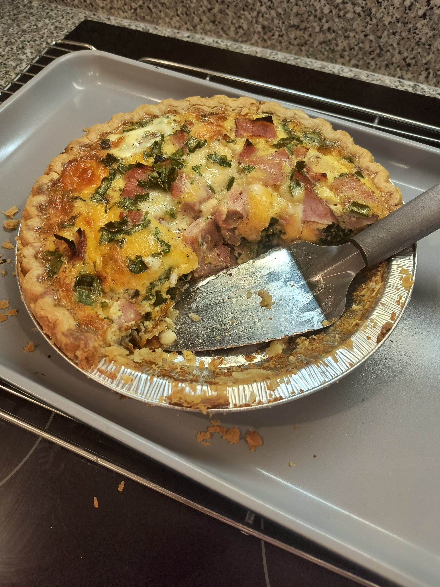 A baked quiche with ham, cheese, and green vegetables sits in a foil pie tin. A slice is missing, and a metal spatula rests inside the quiche on a baking sheet. Crumbs are scattered on the sheet.