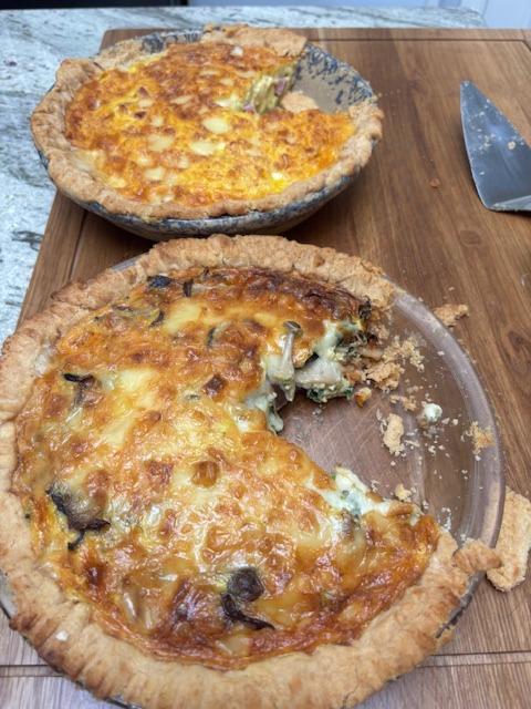 Two homemade quiches with golden, flaky crusts sit on a wooden cutting board. One quiche has a large slice missing, revealing its cheesy, vegetable-filled interior. A knife rests nearby.
