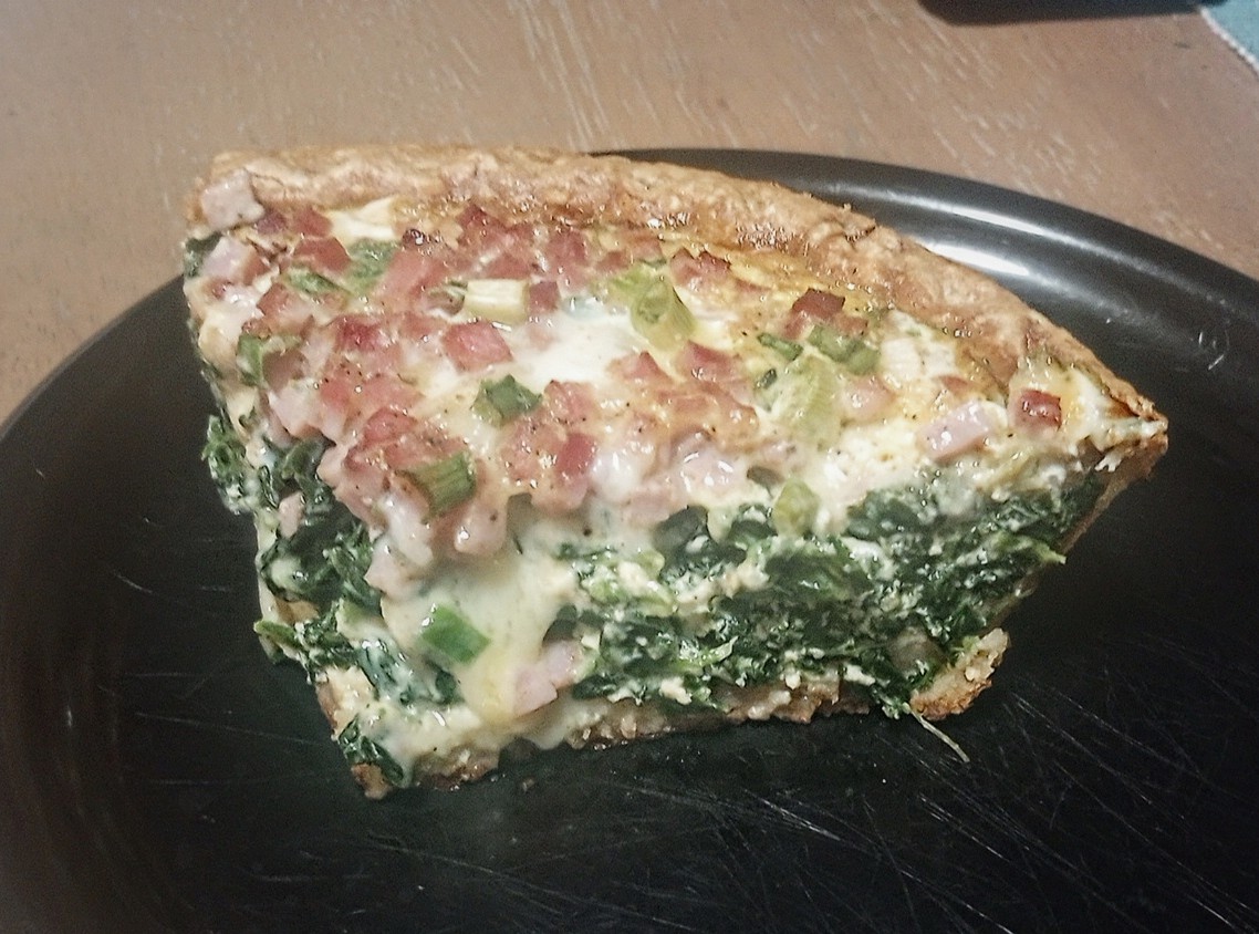 A slice of quiche on a black plate, filled with spinach, diced ham, chopped green onions, and melted cheese, with a golden-brown crust.