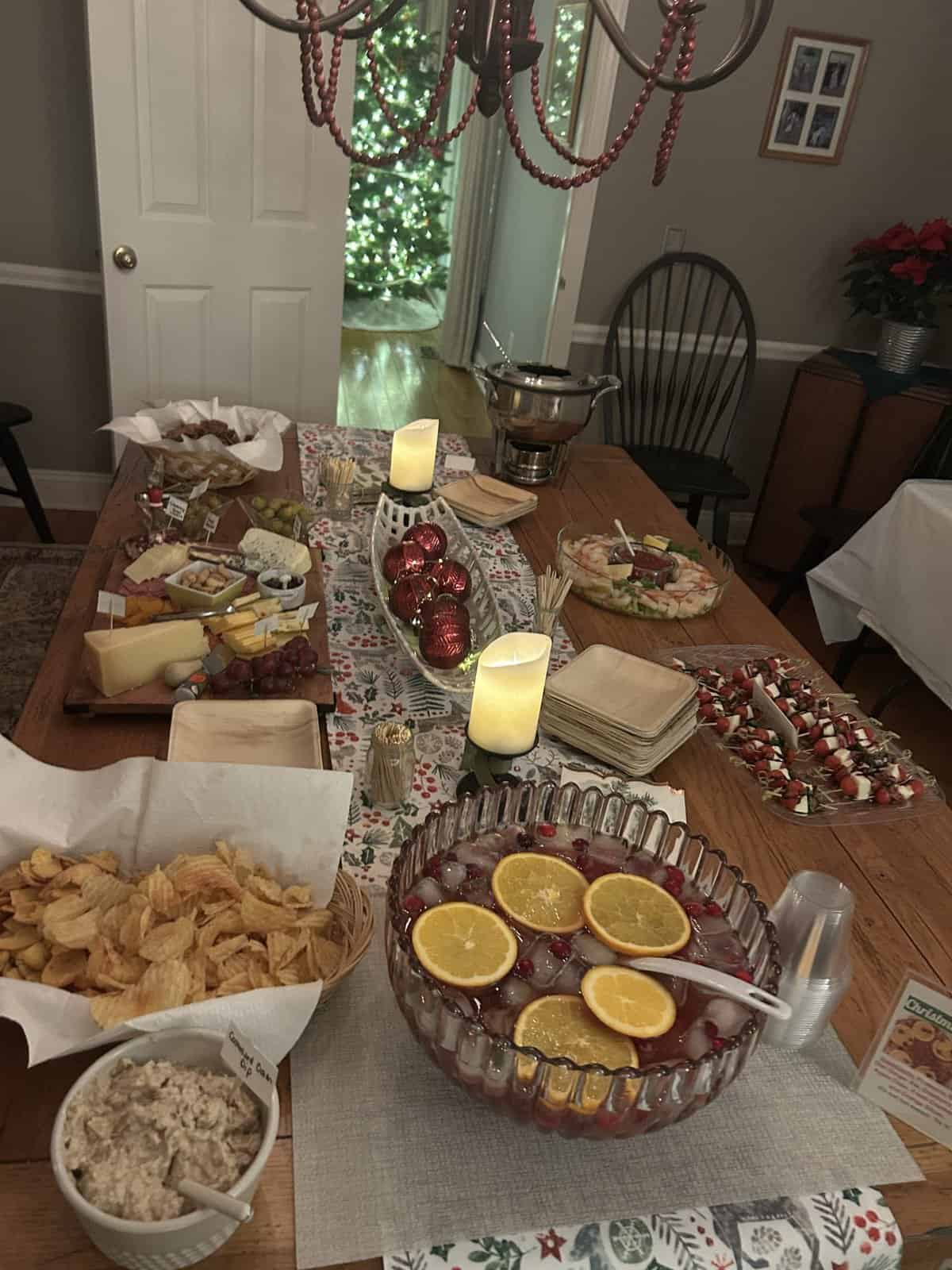 A festive holiday table with snacks, cheese, punch, candles, and a decorated Christmas tree in the background.