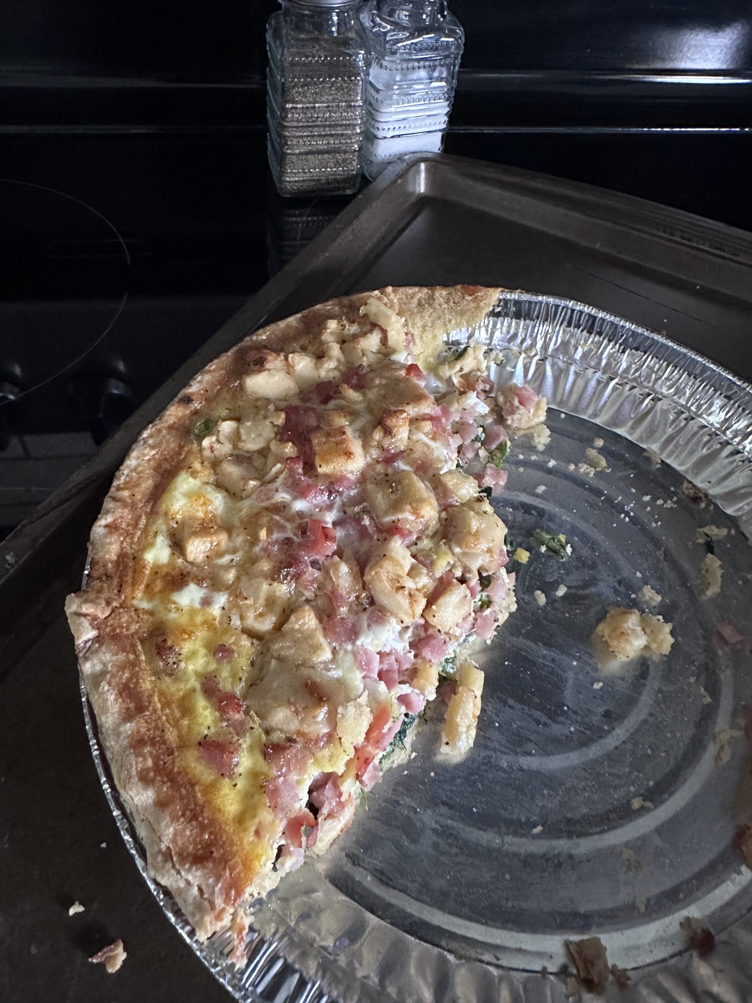 A partially eaten quiche in a foil pie dish sits on a baking sheet. The quiche contains visible pieces of ham and bread cubes. A stack of glass salt and pepper shakers is in the background.