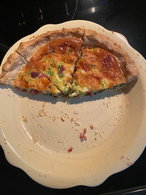 A partially eaten quiche with a golden-brown crust, cut into four slices, sits on a cream-colored pie dish. Some crumbs and bits of filling are scattered on the dish around the quiche.