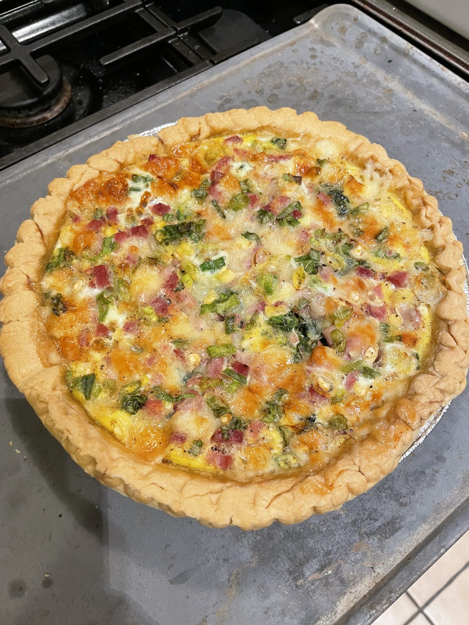A freshly baked quiche with a golden, flaky crust, filled with diced ham, melted cheese, and chopped green herbs, sits on a baking sheet next to a stove.