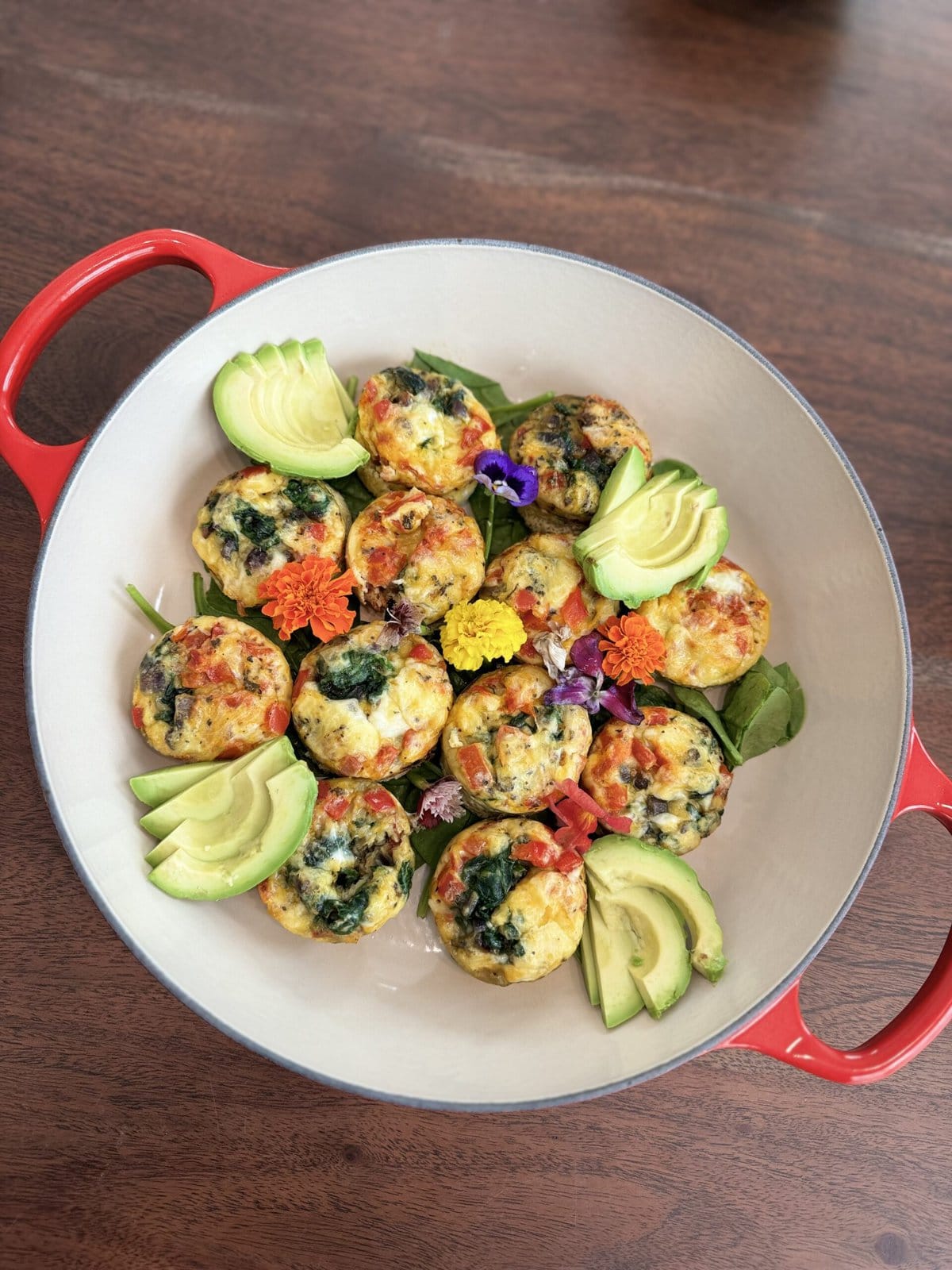 Egg muffins with spinach and veggies, garnished with avocado slices and edible flowers in a red-handled dish.