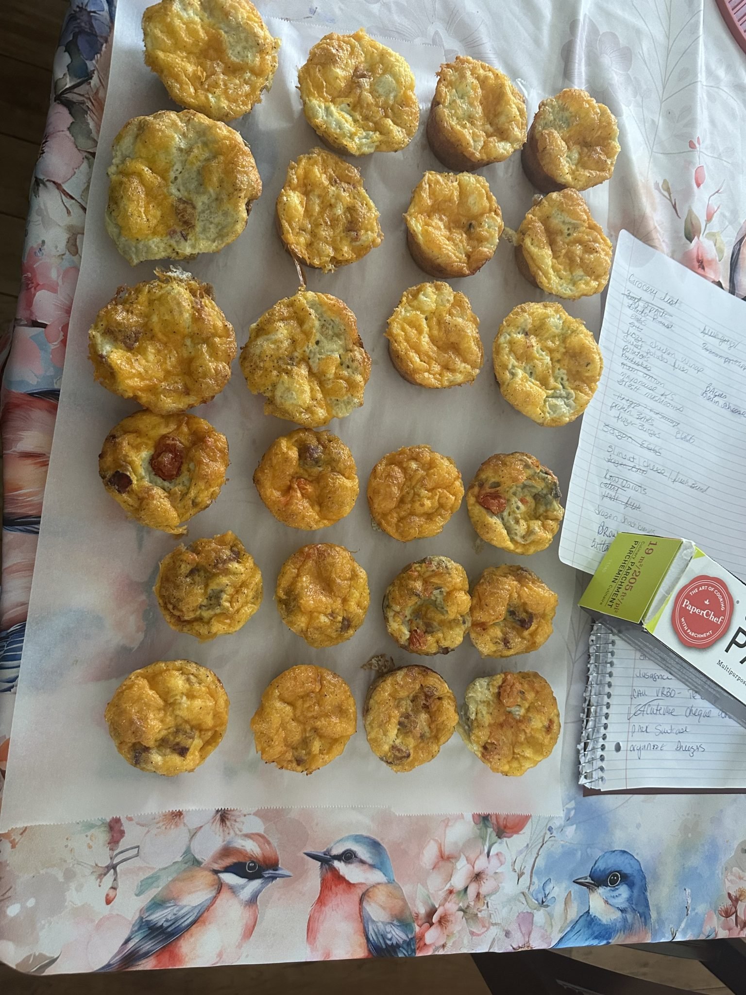 Rows of savory muffins cooling on parchment paper next to a recipe notebook on a bird-patterned tablecloth.