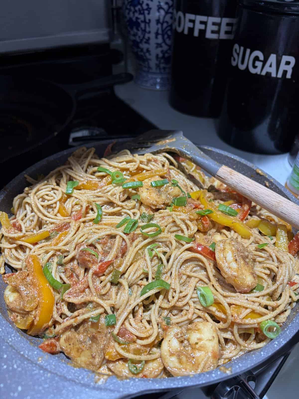 A pan of shrimp pasta with sliced bell peppers and chopped green onions, next to coffee and sugar containers.