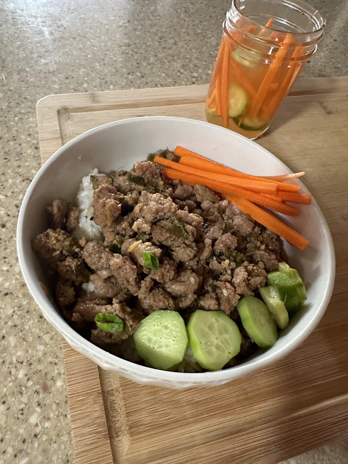 A bowl of rice topped with ground meat, sliced cucumbers, and carrot sticks, with pickled vegetables in a jar nearby.