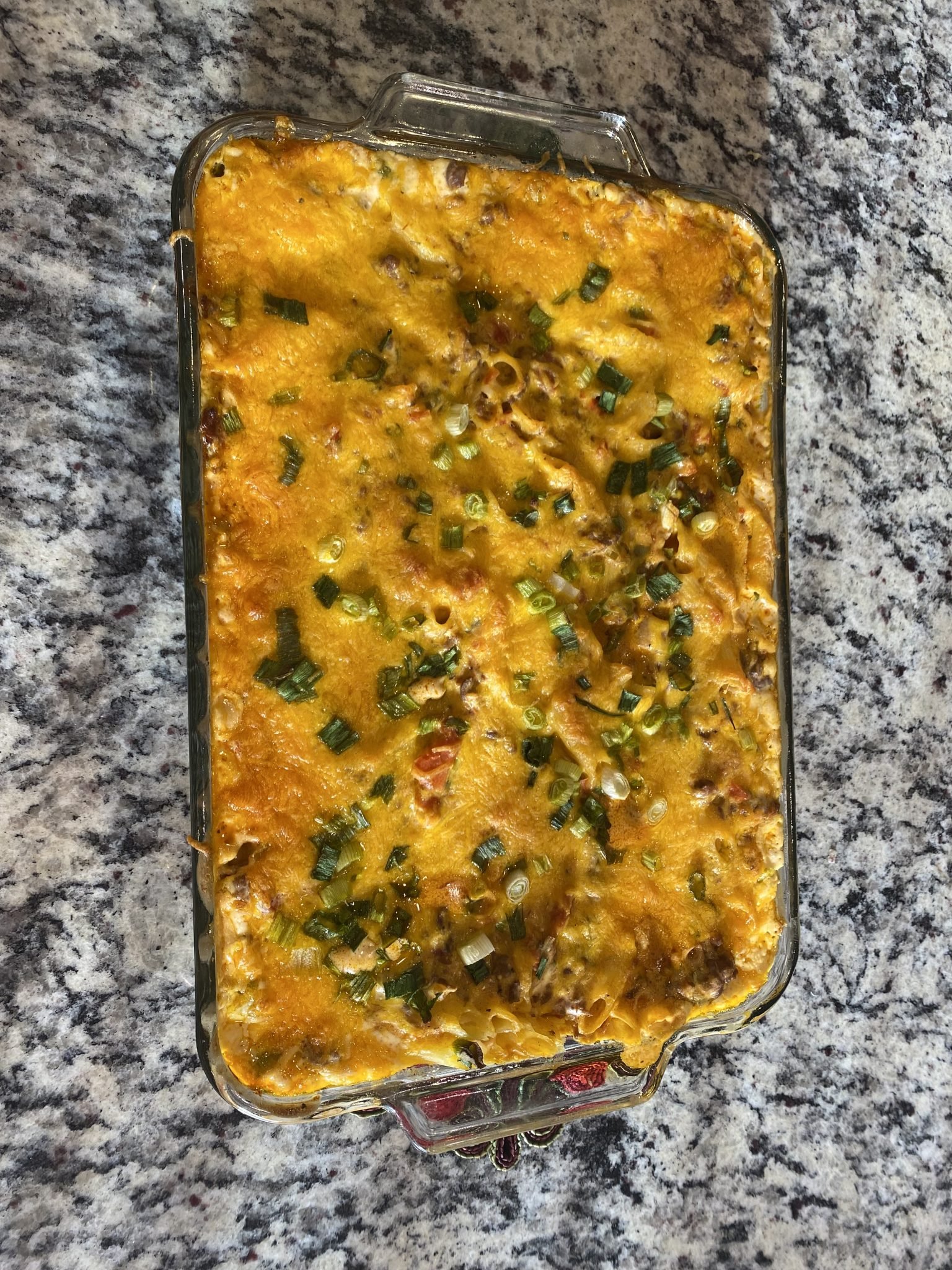 A baked casserole topped with melted cheese and chopped green onions in a glass dish on a granite countertop.