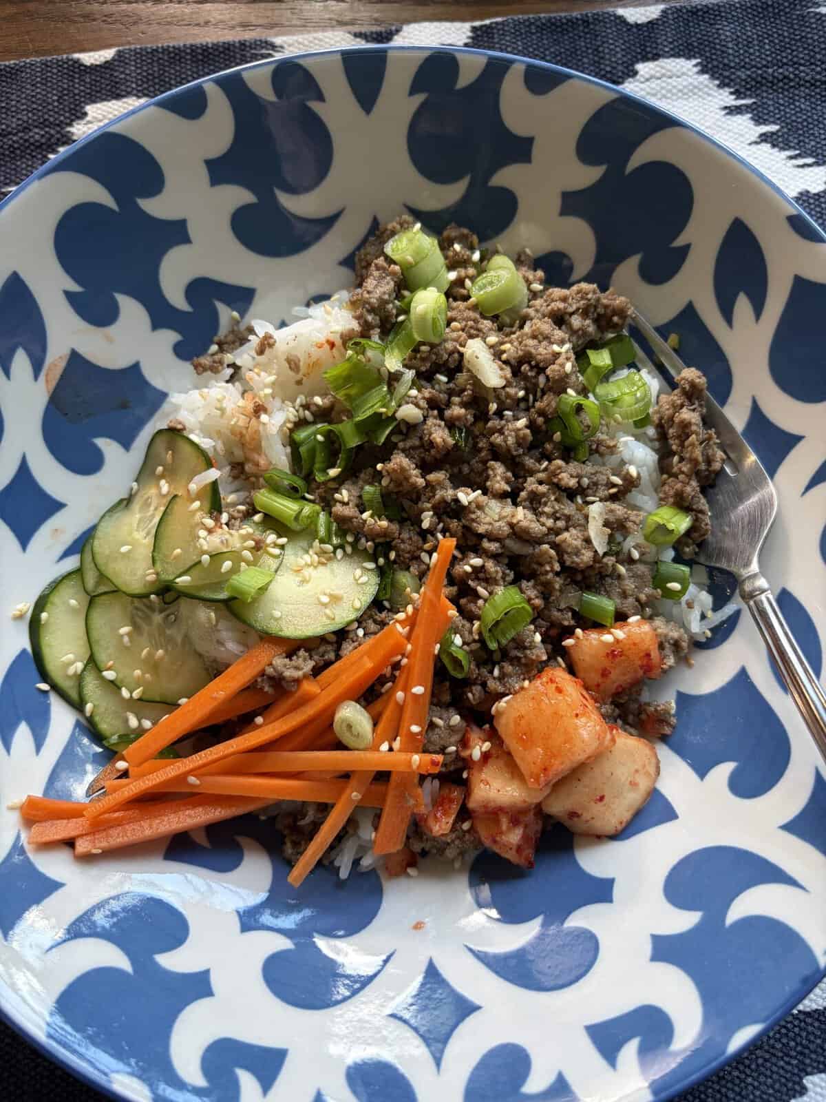 A bowl of rice topped with ground beef, sliced cucumbers, carrots, kimchi, green onions, and sesame seeds.