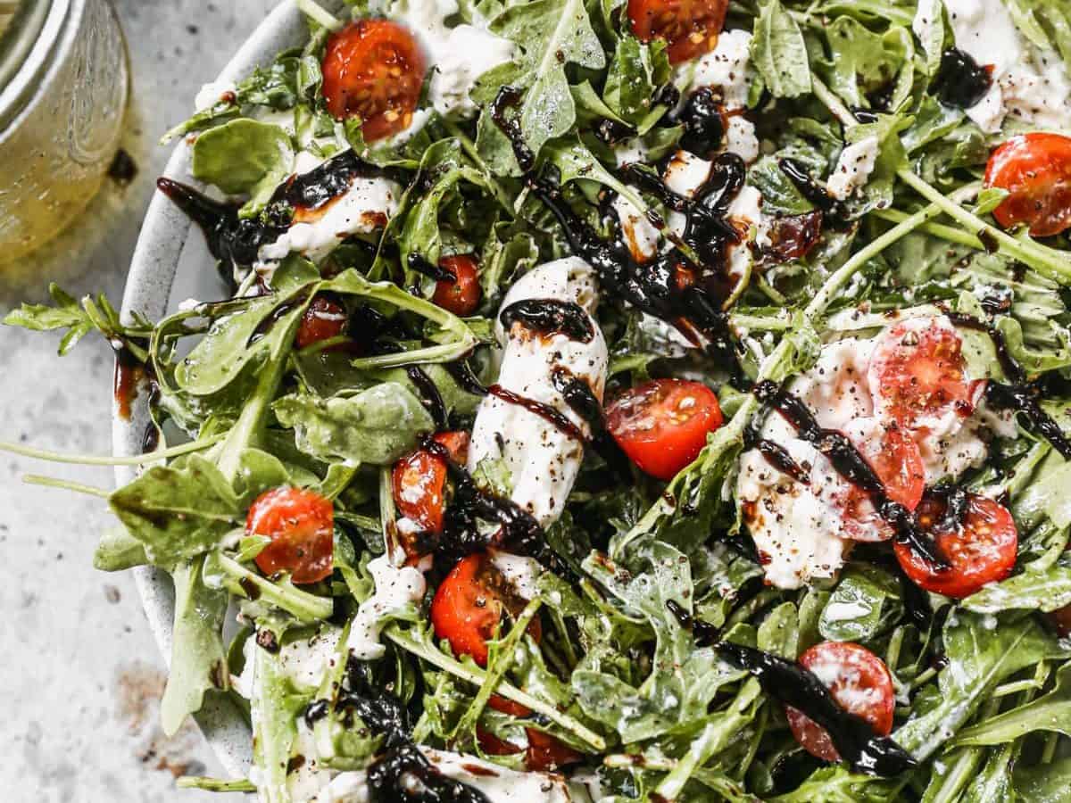 Arugula salad with cherry tomatoes, mozzarella, and balsamic glaze on a white plate.