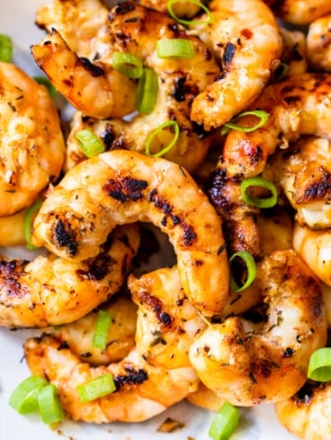 Close-up of grilled shrimp seasoned with herbs and spices, garnished with sliced green onions. The shrimp are slightly charred and arranged on a white plate.