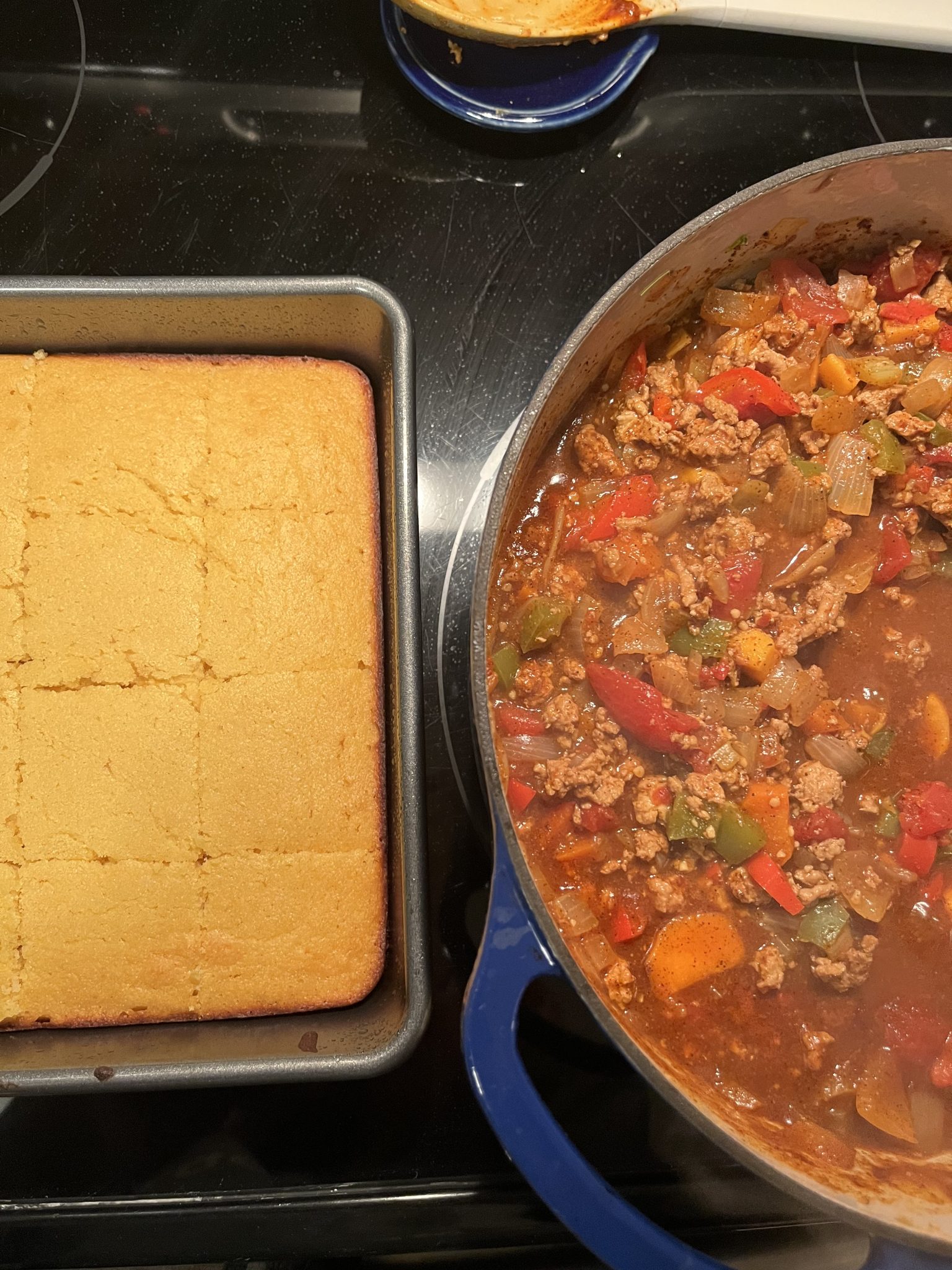 A pan of baked cornbread is on the left, and to its right is a blue pot filled with chunky chili containing ground meat, tomatoes, peppers, and onions. Both are on a black stovetop.