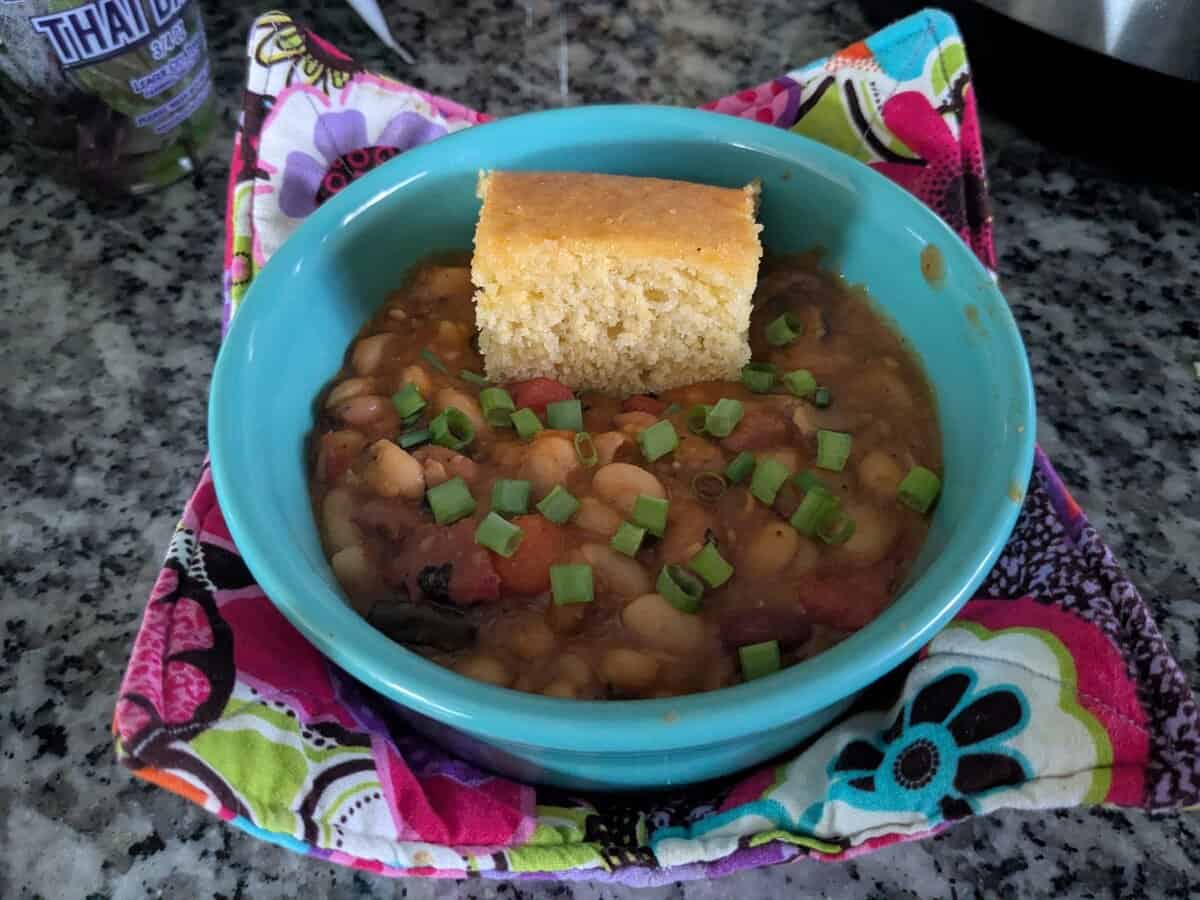 A bowl of bean soup topped with chopped green onions and a slice of cornbread, set on a colorful cloth.