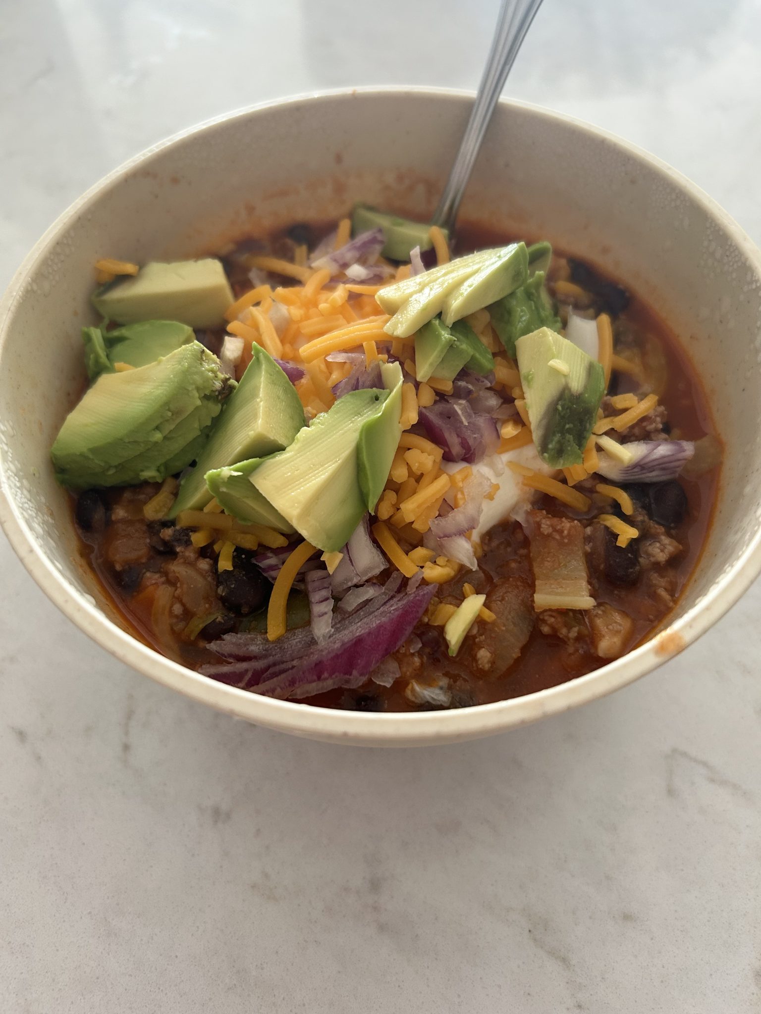 A bowl of chili topped with sliced avocado, shredded cheddar cheese, diced red onions, and a dollop of sour cream. A spoon is visible in the bowl, set on a light-colored surface.