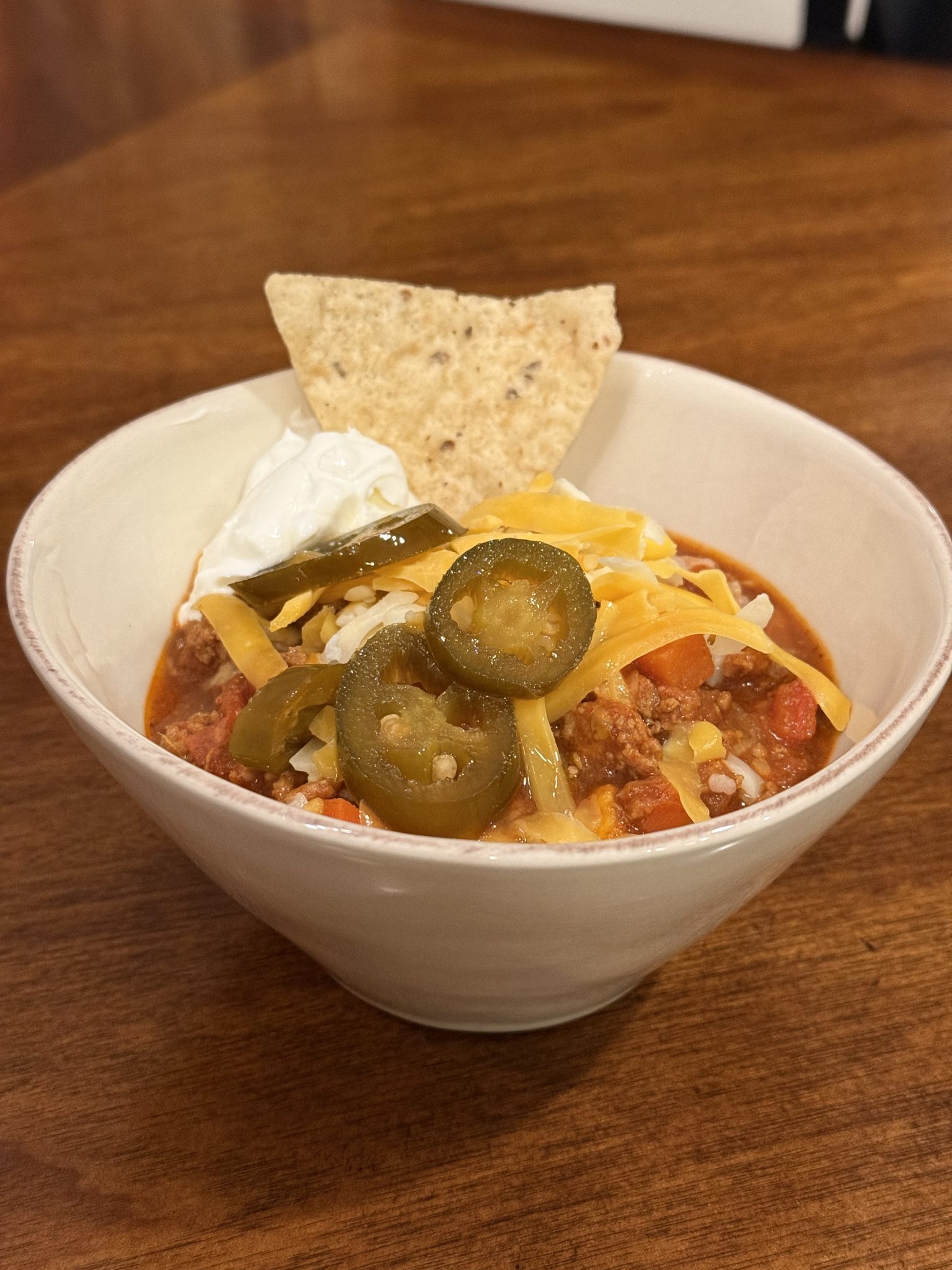 A bowl of chili topped with shredded cheese, jalapeños, sour cream, and a tortilla chip on a wooden table.