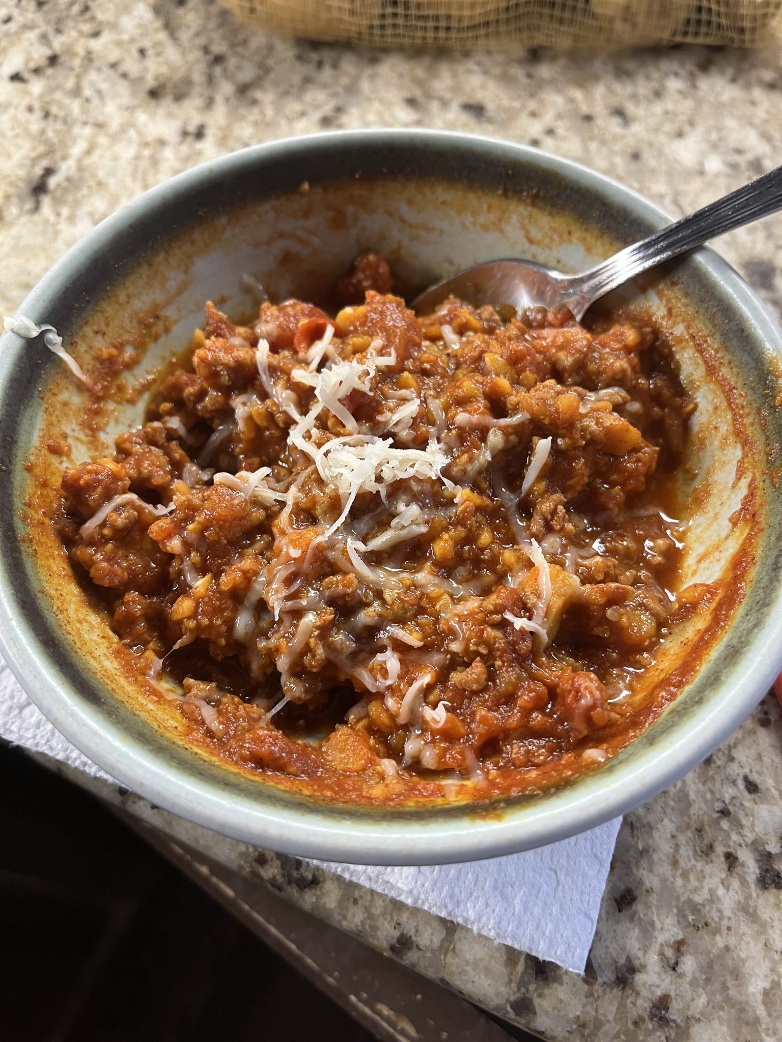 A bowl of hearty chili topped with shredded cheese sits on a countertop. The chili is thick with a tomato-based sauce, beans, and ground meat mixed in. A spoon rests in the bowl. The bowl is placed on a paper towel.
