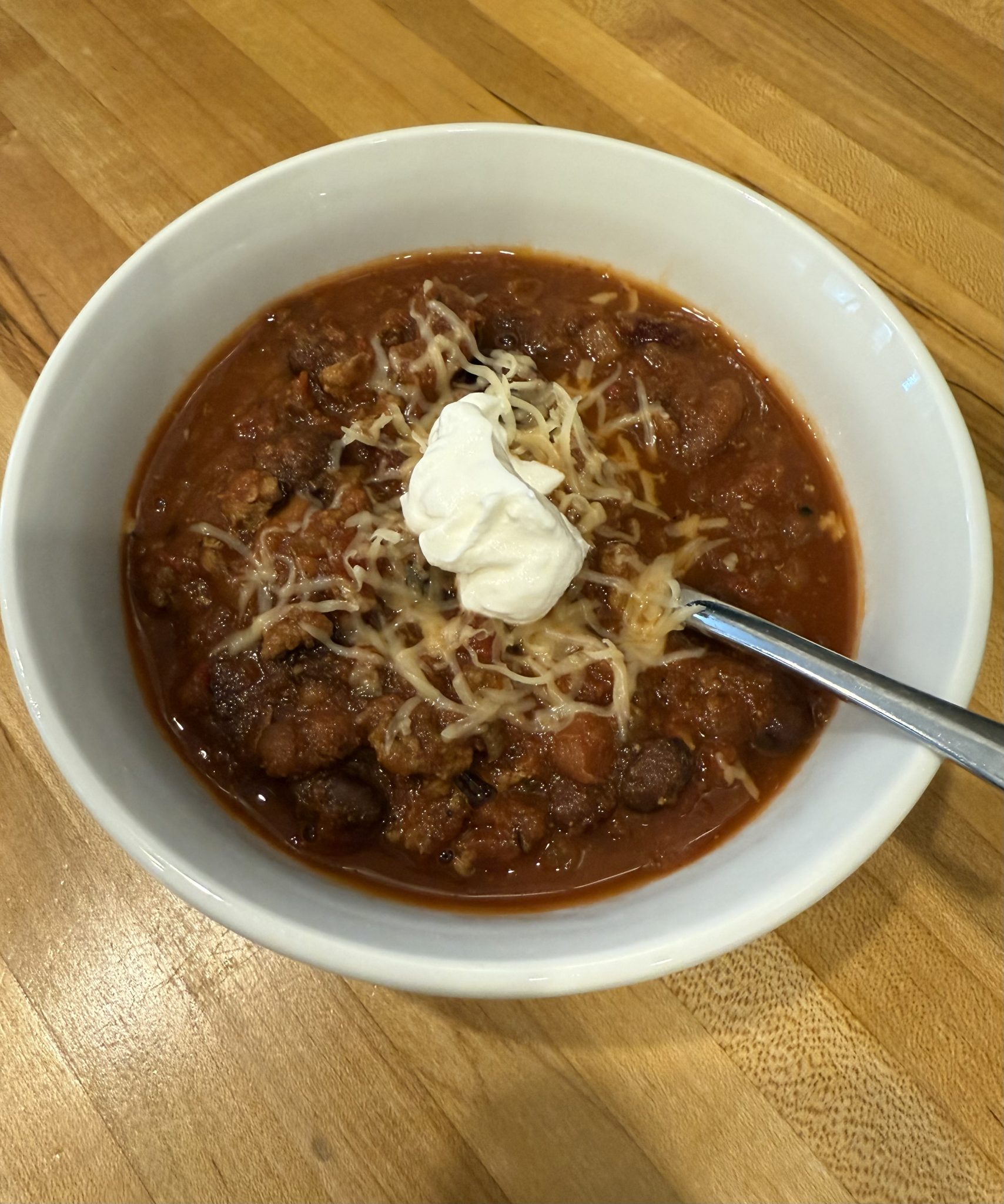 A bowl of chili with ground meat and beans, topped with shredded cheese and a dollop of sour cream. A spoon is resting in the bowl, and the dish is placed on a wooden surface.