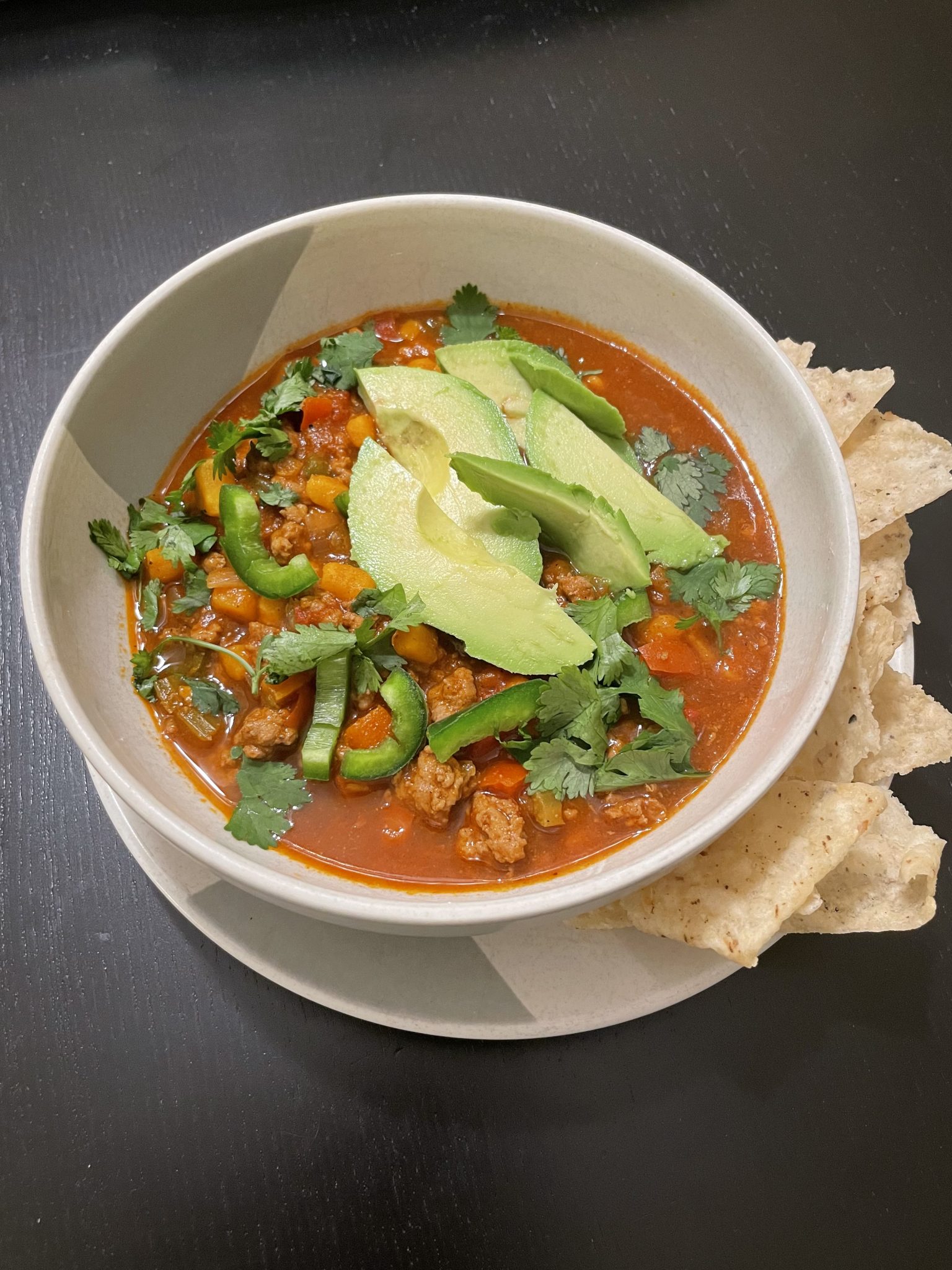 A bowl of chili topped with sliced avocado, jalapeños, and cilantro is served with tortilla chips on the side. The chili includes visible chunks of meat, tomatoes, and bell peppers, set against a dark table surface.