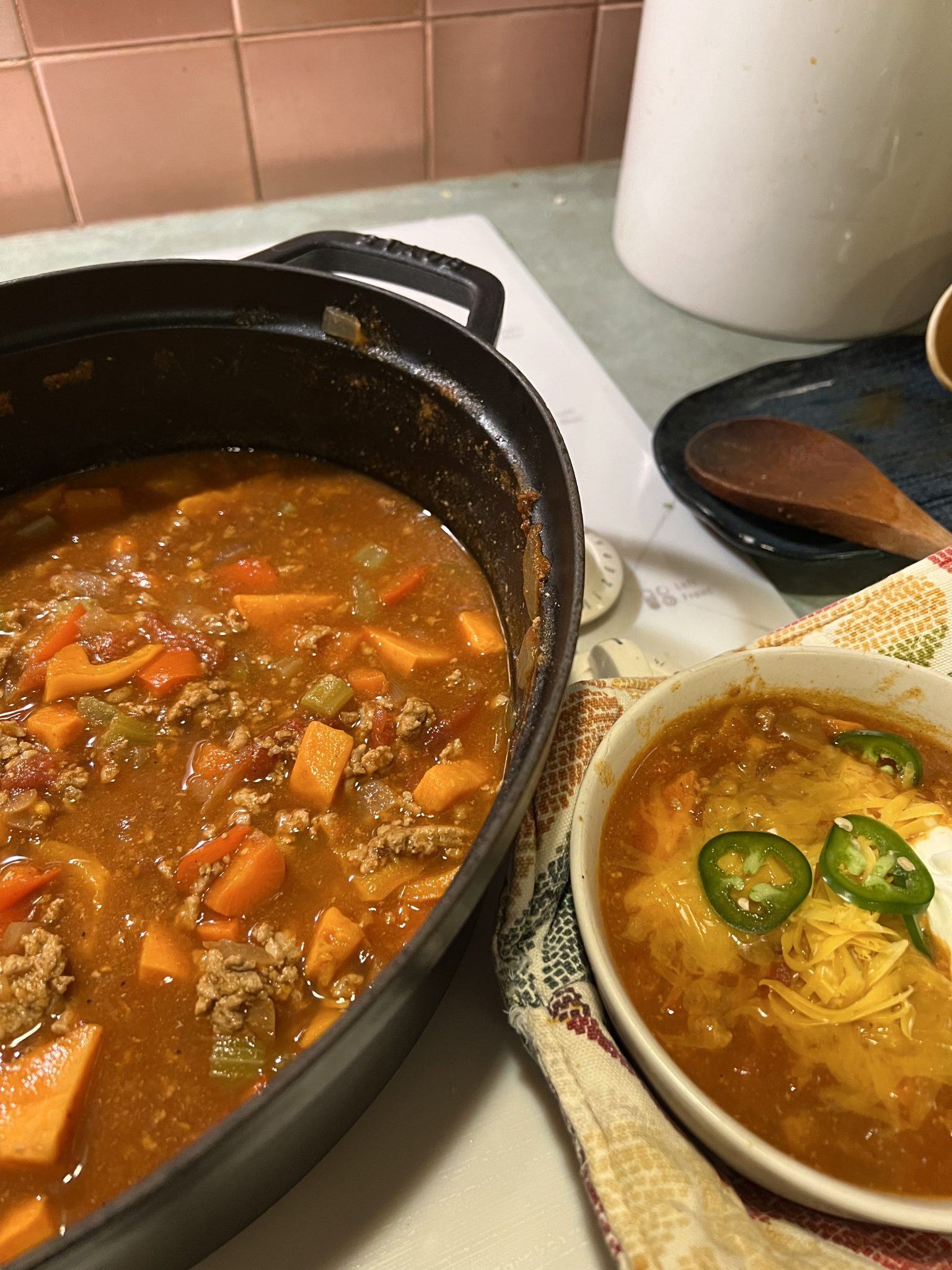 A pot of hearty chili with beef, carrots, and celery next to a bowl filled with chili, topped with shredded cheese, sour cream, and sliced jalapeños. A wooden spoon rests on a cloth nearby.