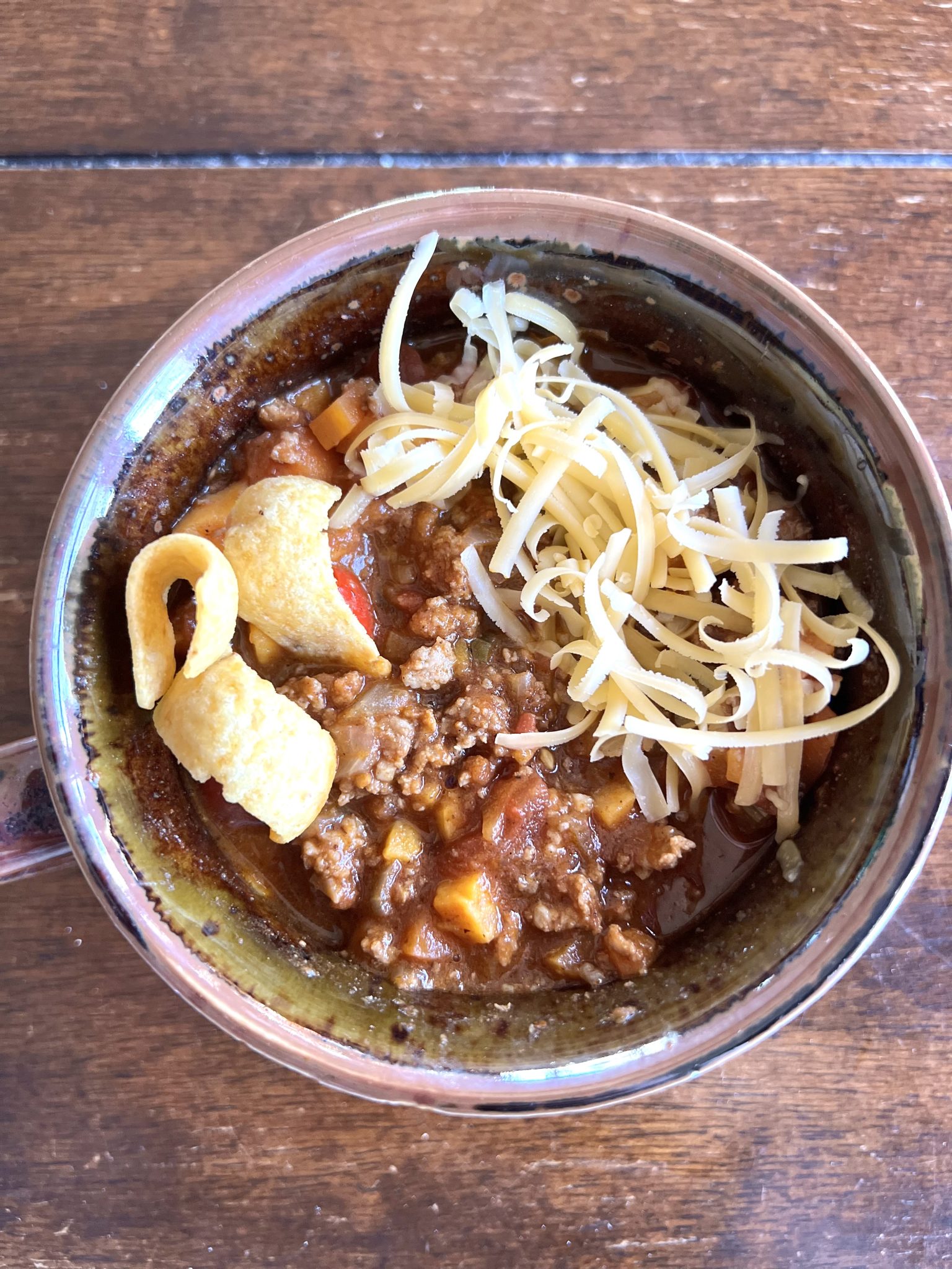A ceramic bowl brims with turkey chili, topped with shredded cheese and corn chips, all placed on a rustic wooden table. Tender pieces of meat and vibrant vegetables peek through for a hearty delight.