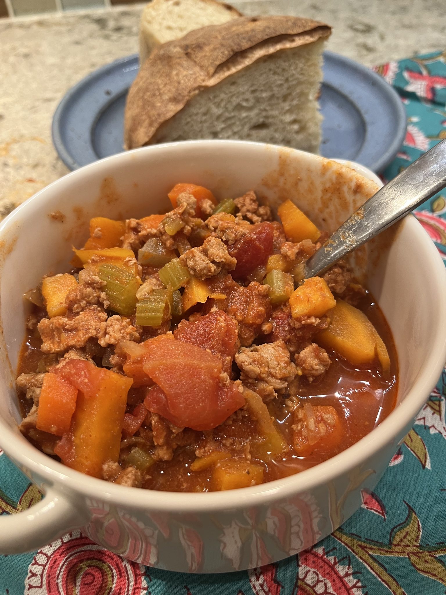 A bowl of hearty stew with ground meat, tomatoes, carrots, celery, and other vegetables. A spoon is resting inside the bowl. In the background, there are two slices of bread on a plate. The tablecloth is colorful and patterned.
